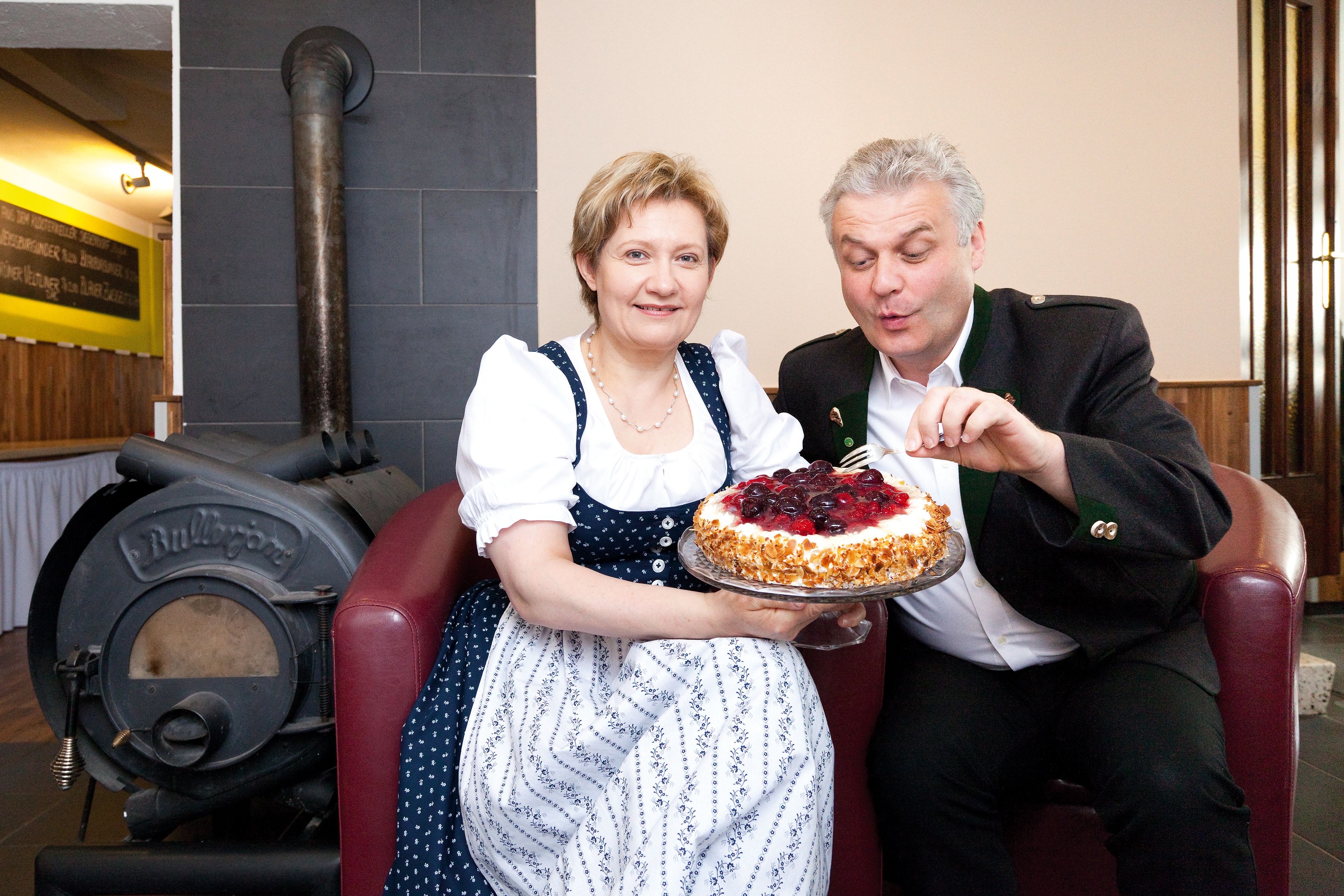 A man and a woman in traditional dress hold a cake in front of an oven.