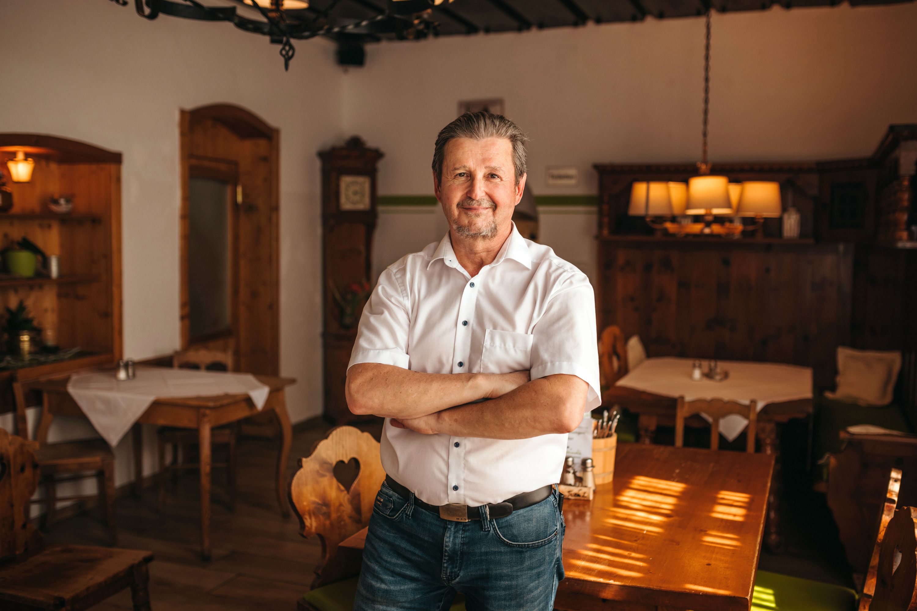 A man in a traditional dining room with wooden furniture, dressed in a white shirt and jeans, stands with his arms folded and smiles.