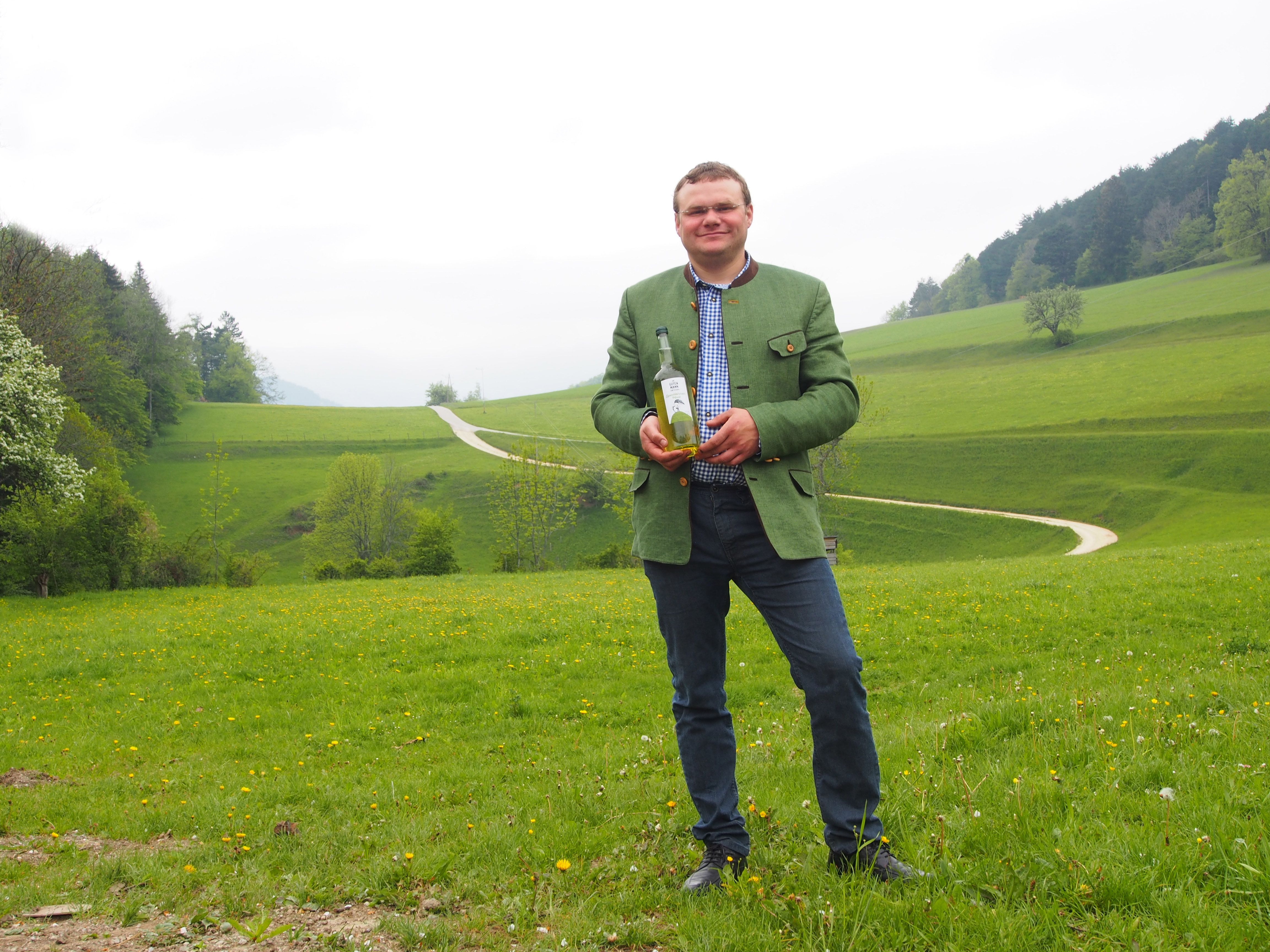 Man in green jacket holding a bottle on a green meadow with hills in the background.