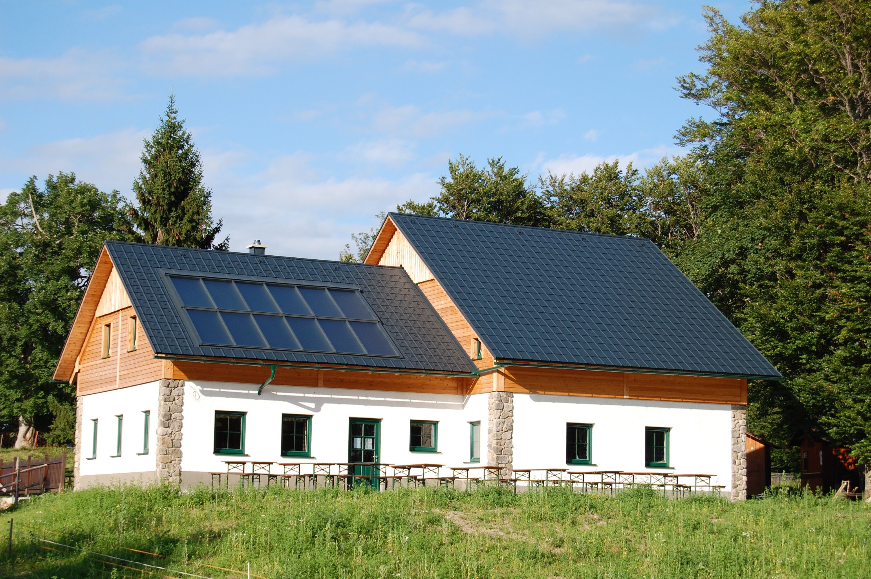 A hut with solar panels on the roof, surrounded by trees and meadows.