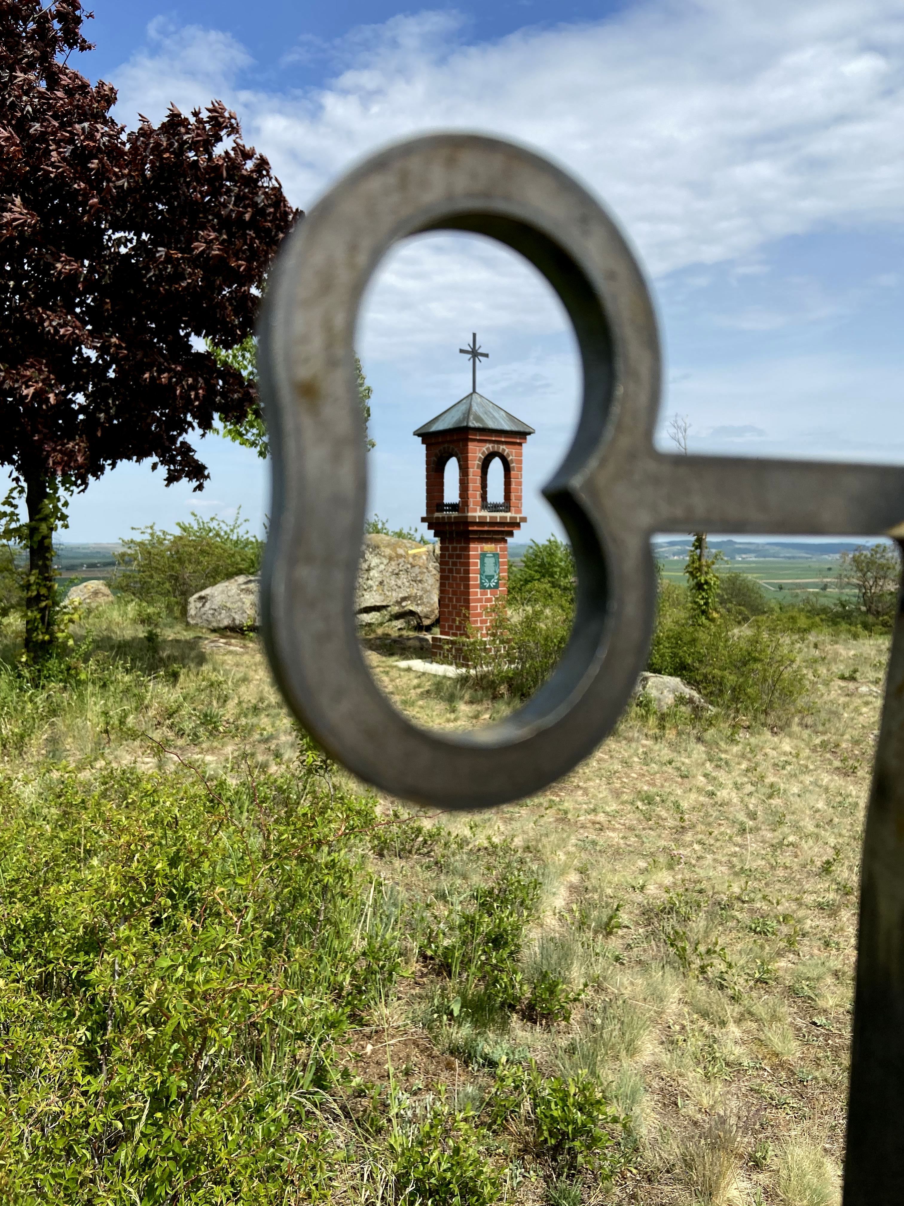 A small brick chapel with a cross on a hill, photographed through a heart-shaped metal grid.
