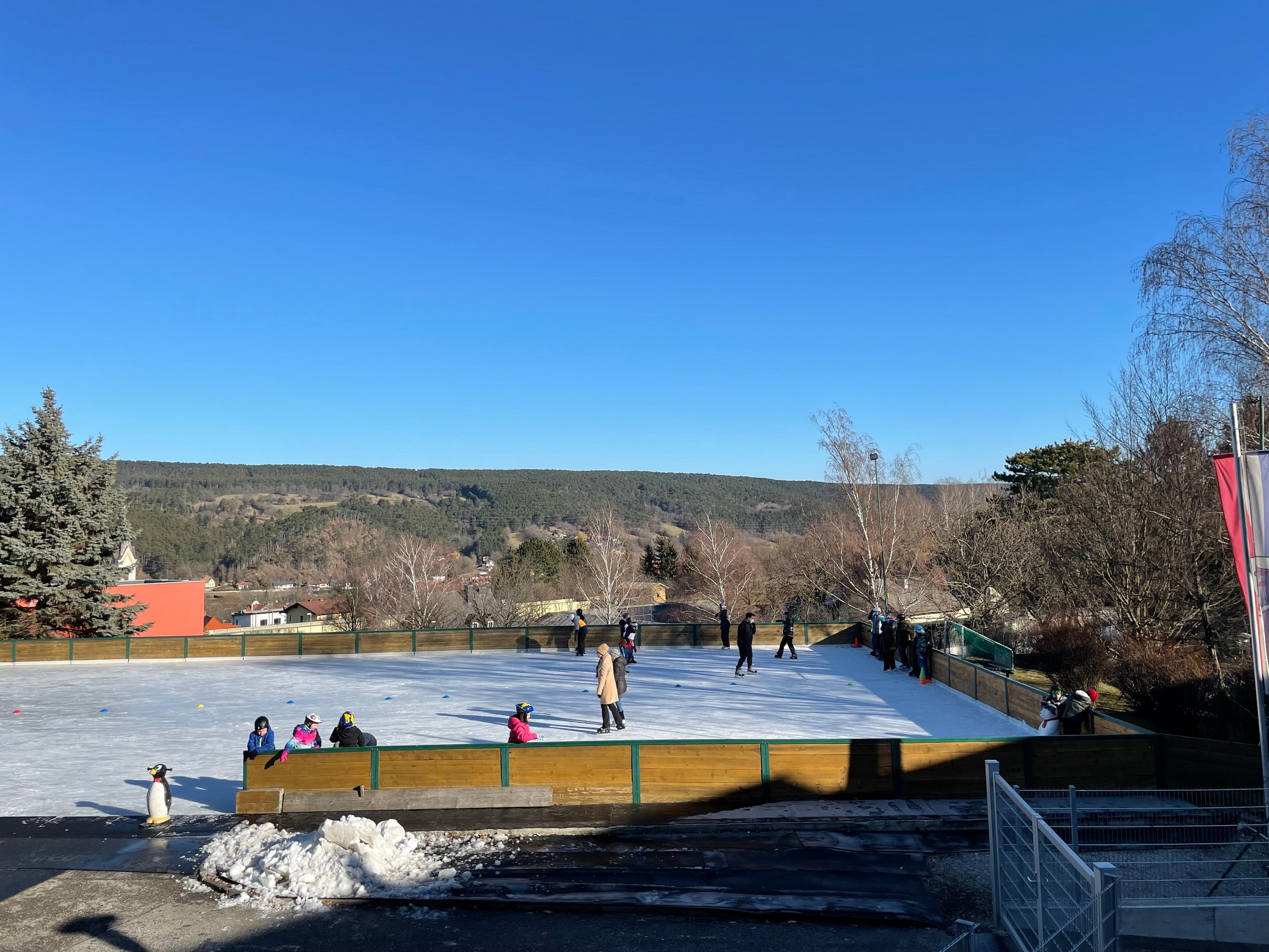 Ice rink with people and a penguin figure, surrounded by trees and hills under a blue sky.