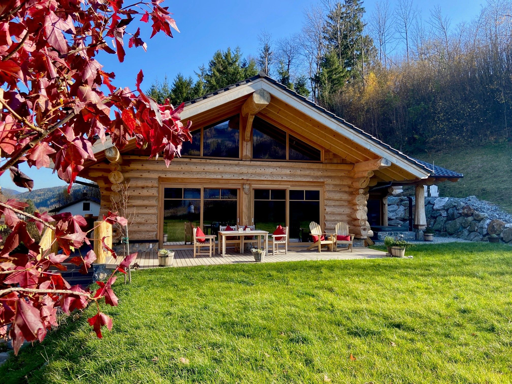 A log cabin with a large terrace, surrounded by green meadows and autumnal trees.