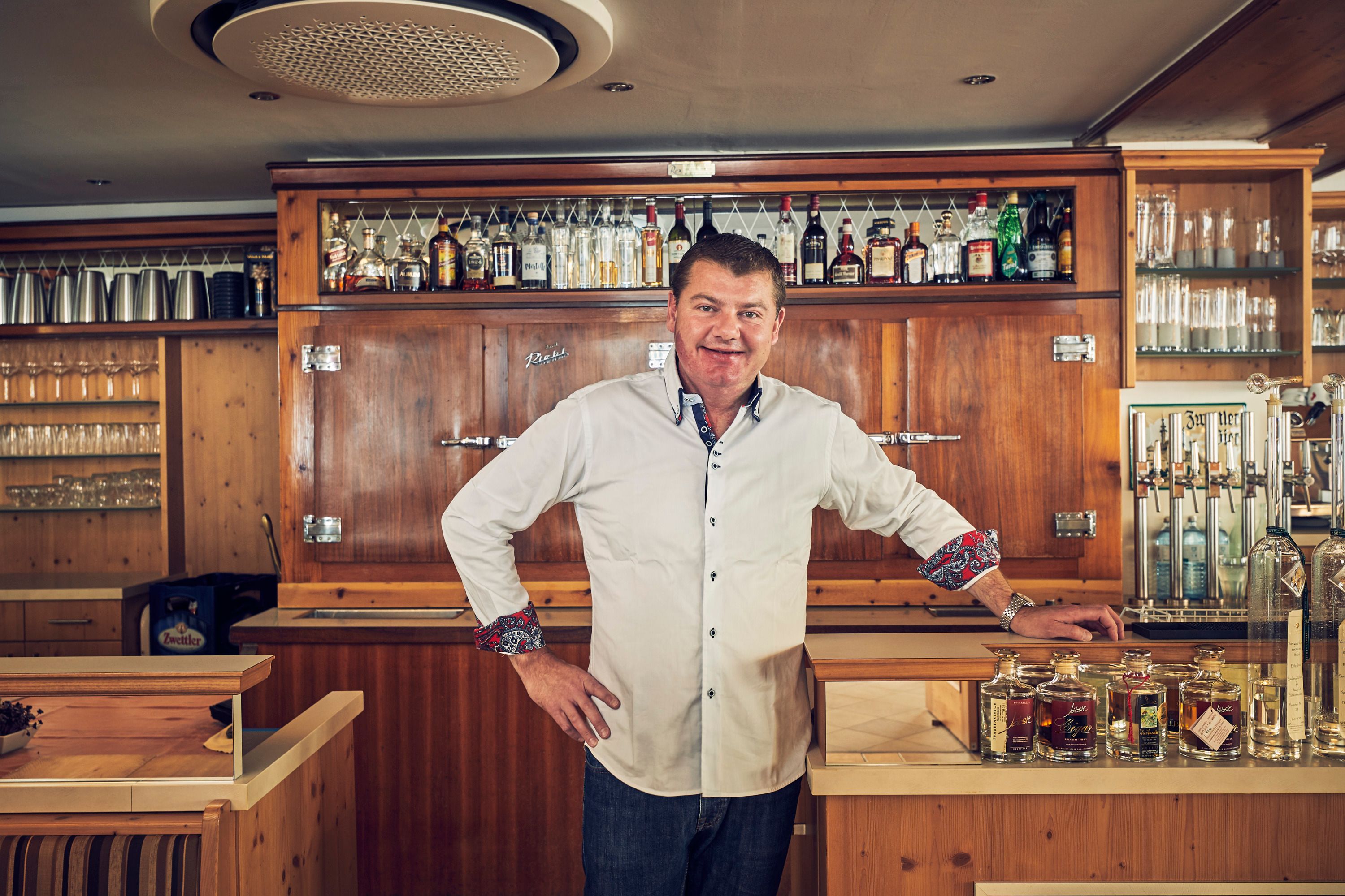 A man in a white shirt stands behind a bar with bottles and glasses in the background.