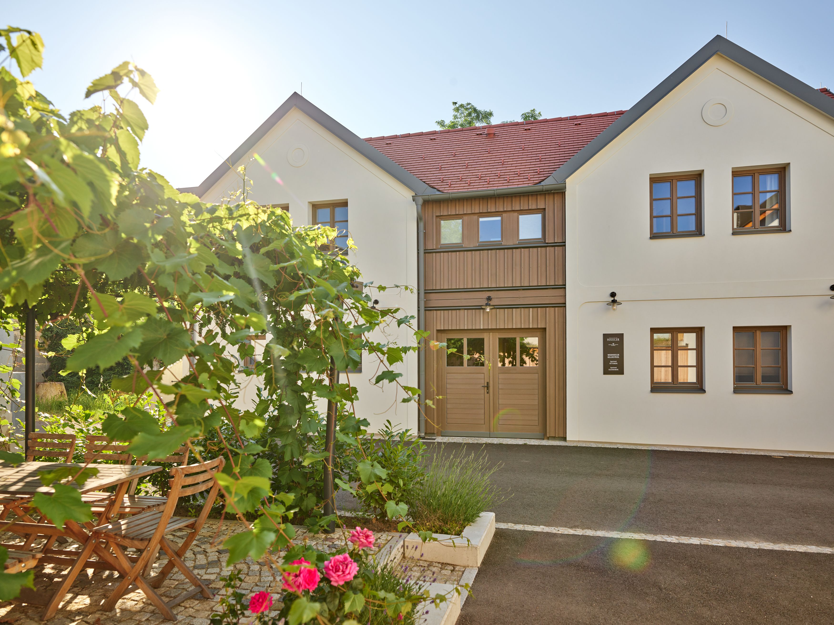 A modern, two-storey building with white walls and red roof tiles. In the foreground, a garden with a wooden table, chairs and flowering plants.