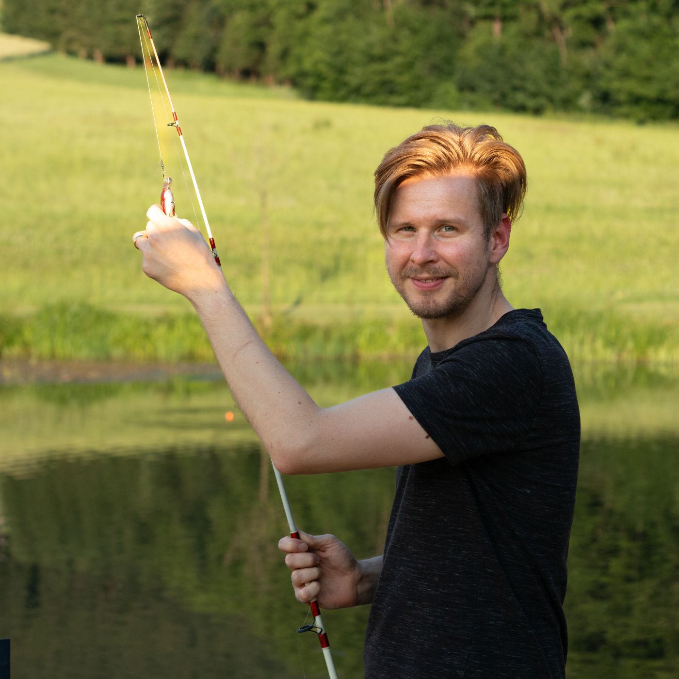 Man with fishing rod at a pond in front of a green landscape.
