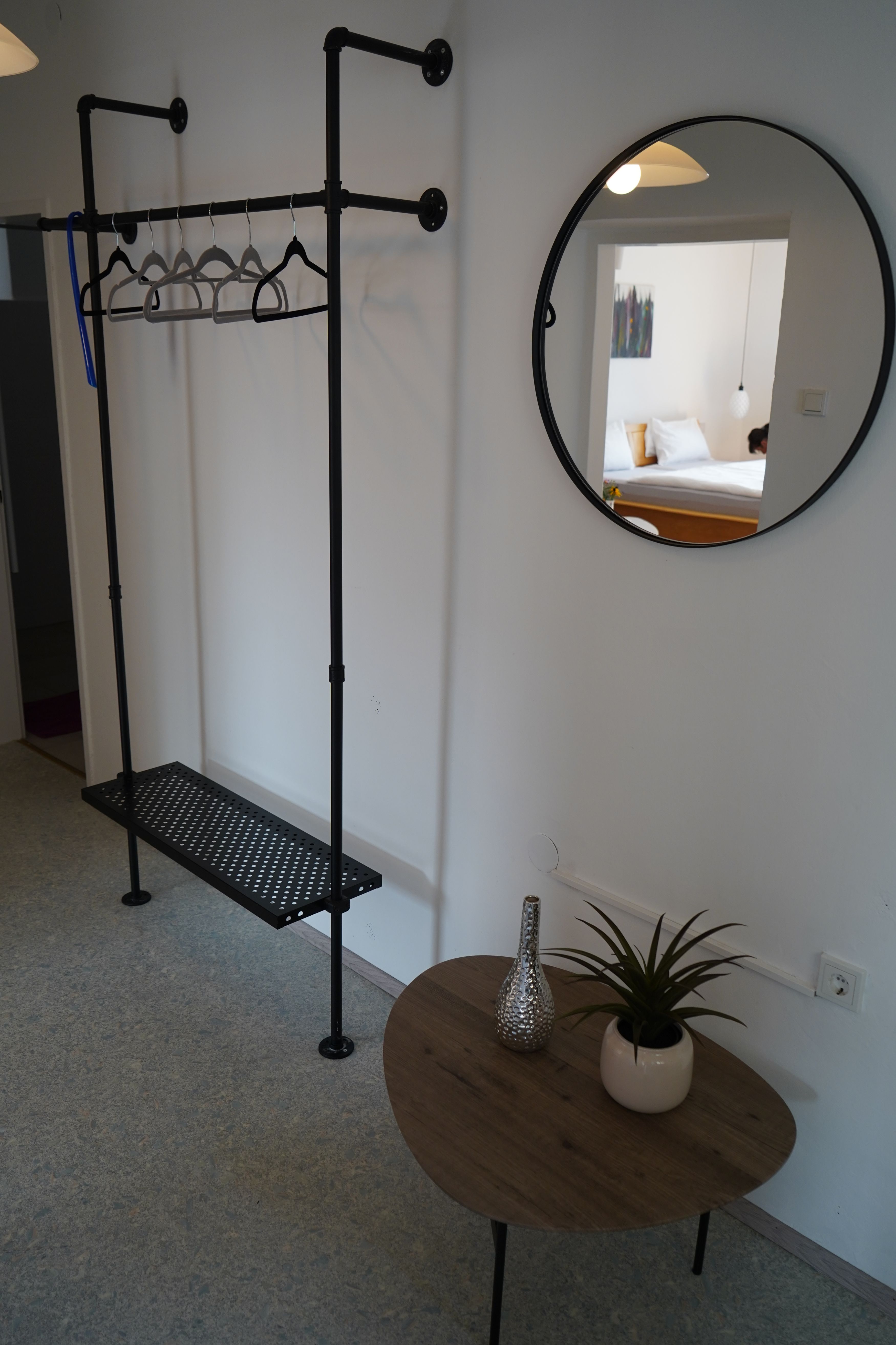 Interior view of a room with clothes rack, round mirror and small table with plant and vase.