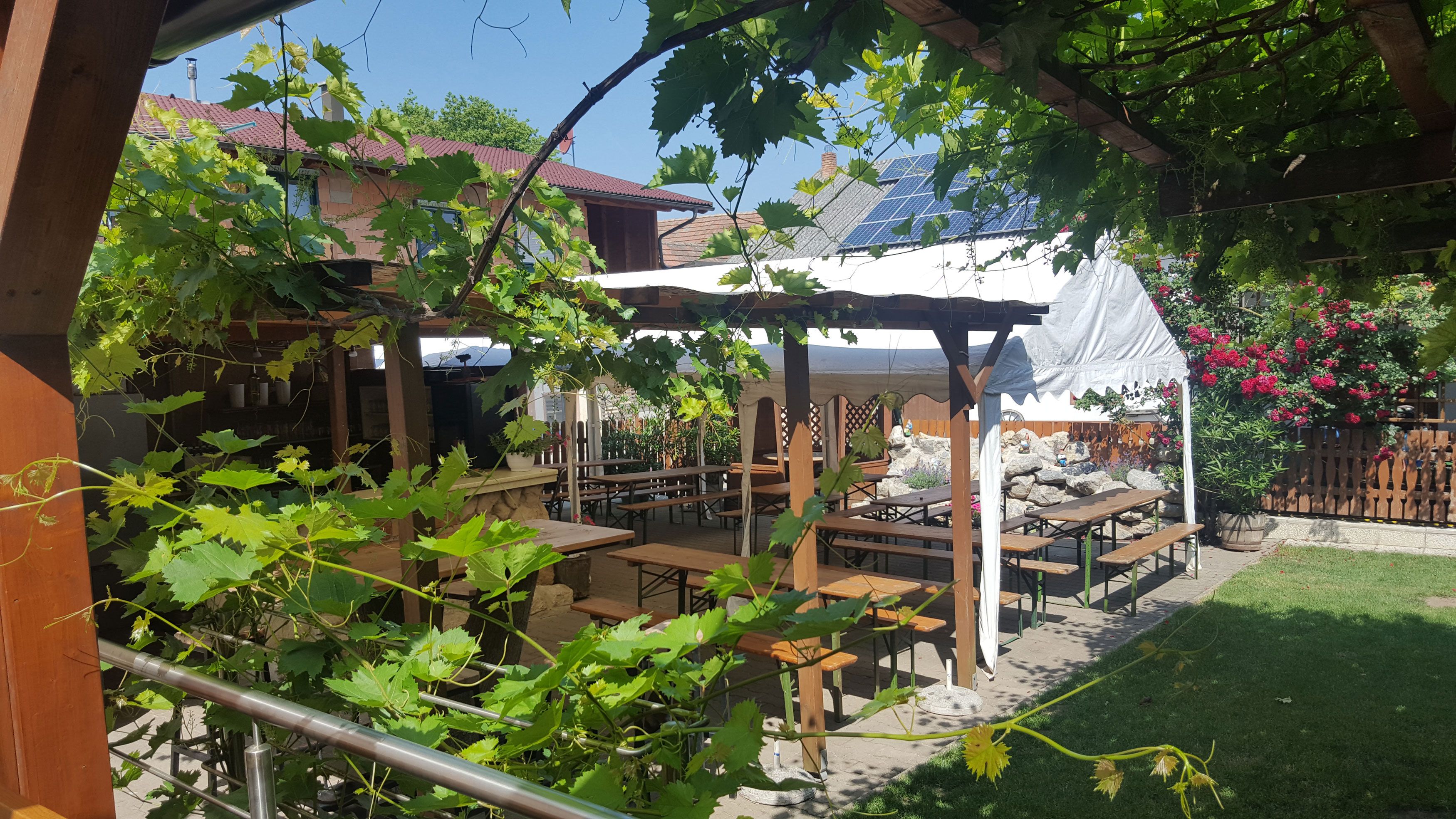 A cozy outdoor area with wooden tables and benches under a leafy canopy, surrounded by plants and flowers.