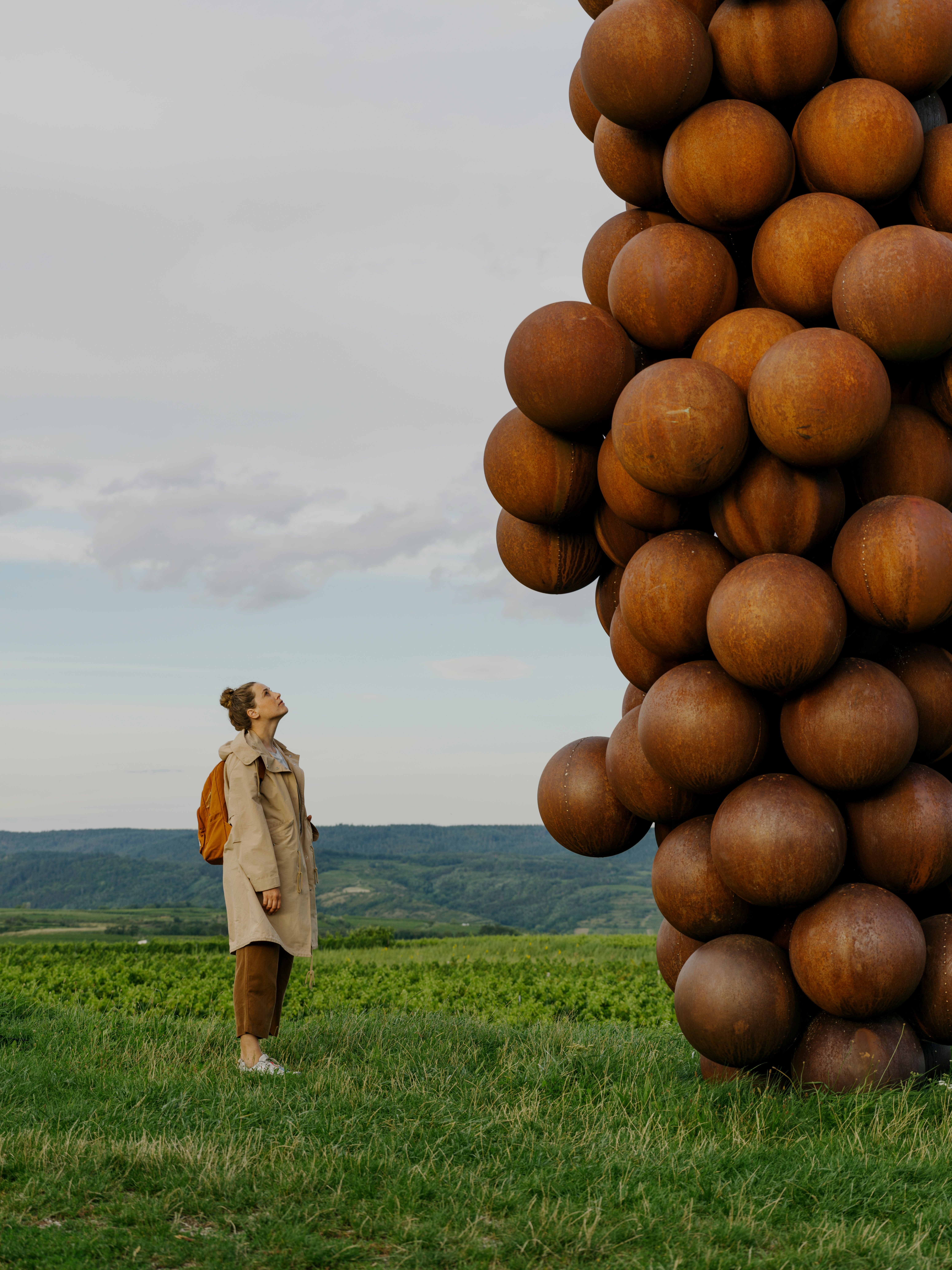 Two people walk past a large sculpture made of spherical elements, surrounded by vineyards.