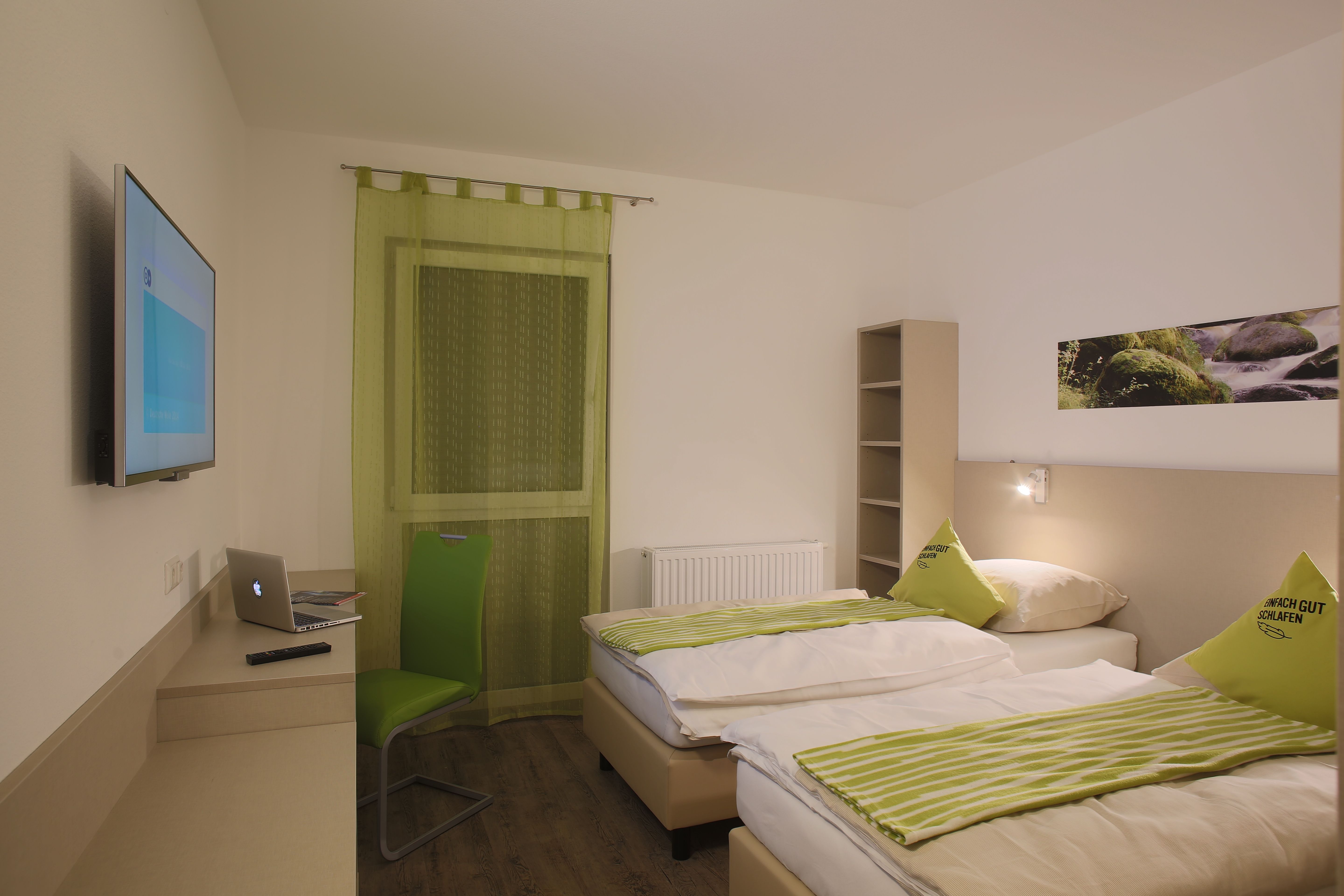 Modern motel room with two beds, green chair, laptop on table and wall-mounted TV.