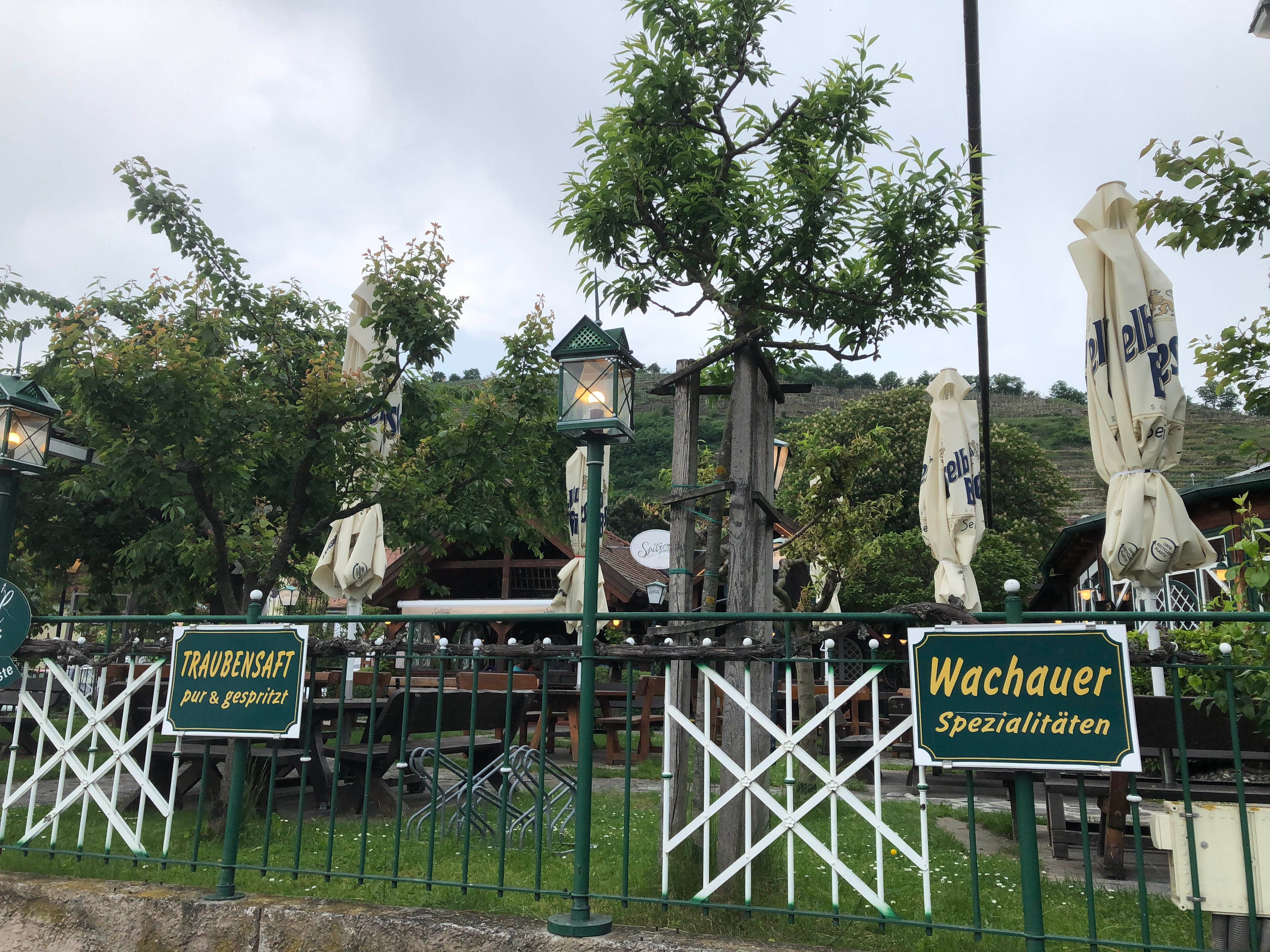 Guest garden with signs for grape juice and Wachau specialties, surrounded by trees and closed parasols.
