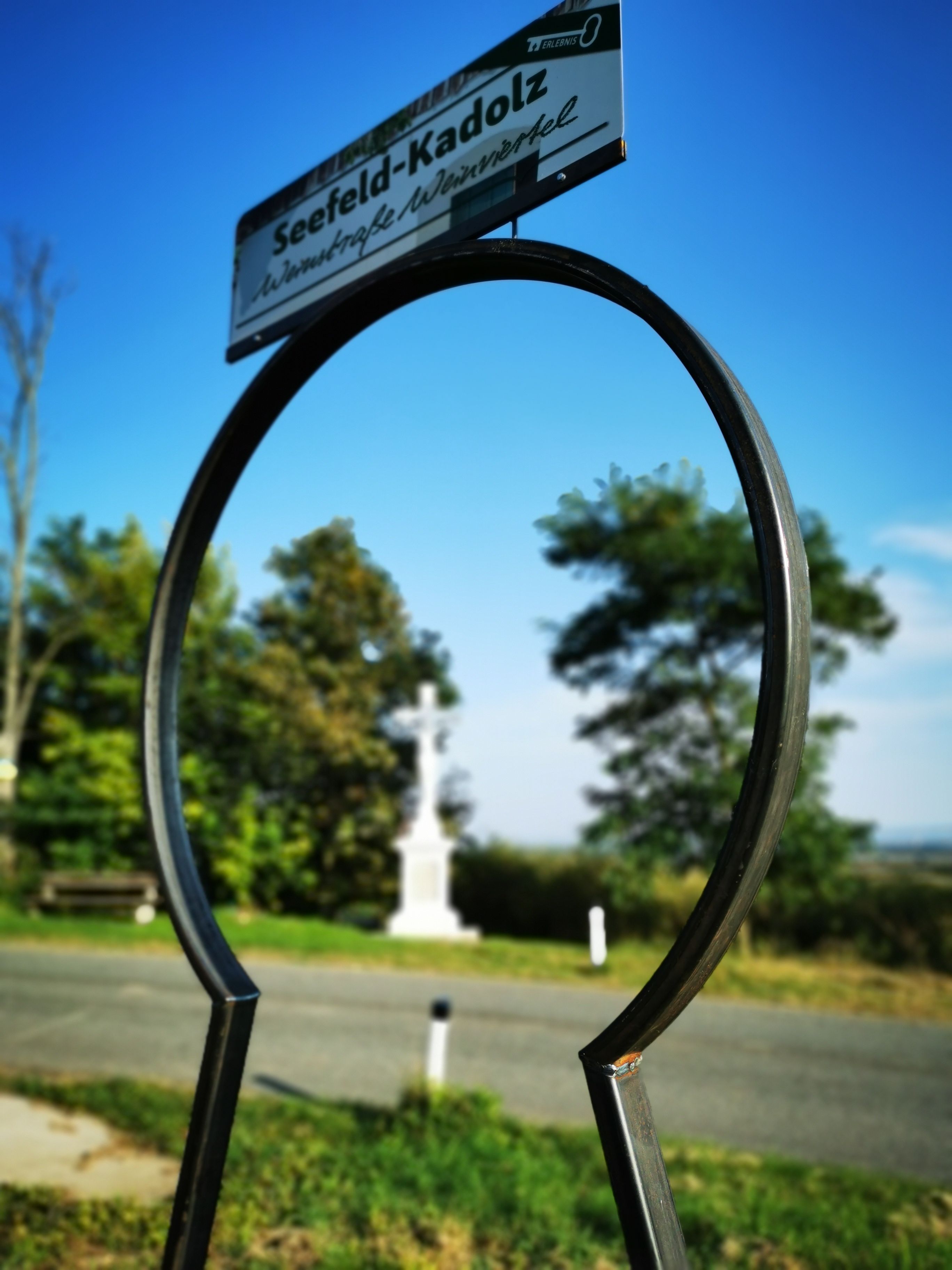 A sign with the inscription 'Seefeld-Kadolz' against a blue sky, with a white cross and trees in the background.