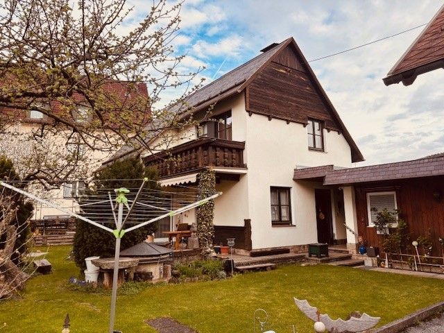 A two-storey house with a balcony and garden, surrounded by trees and a tumble dryer in the foreground.