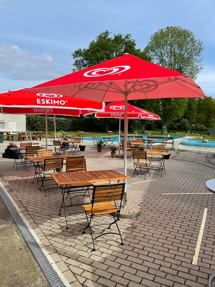 An outdoor pool with red parasols and empty wooden tables on a paved terrace.