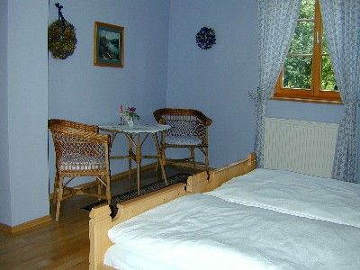 A cozy room with two beds, a small table and two chairs. Windows with curtains and wall decorations are visible.
