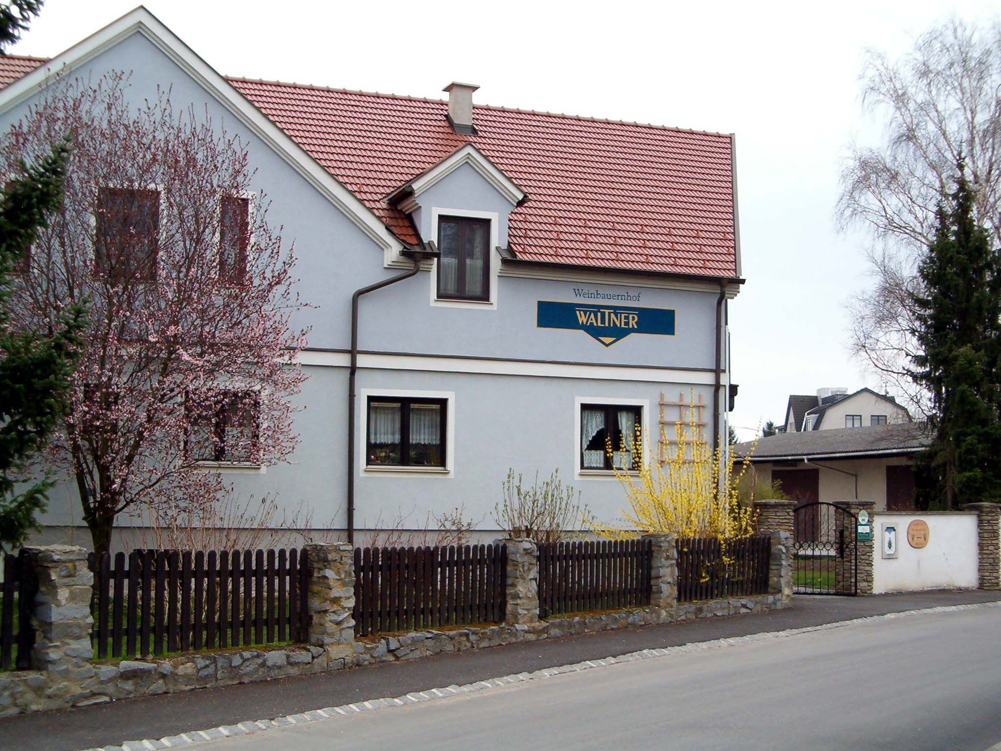 A two-storey house with a red roof and blue façade, surrounded by blossoming trees and a wooden fence.