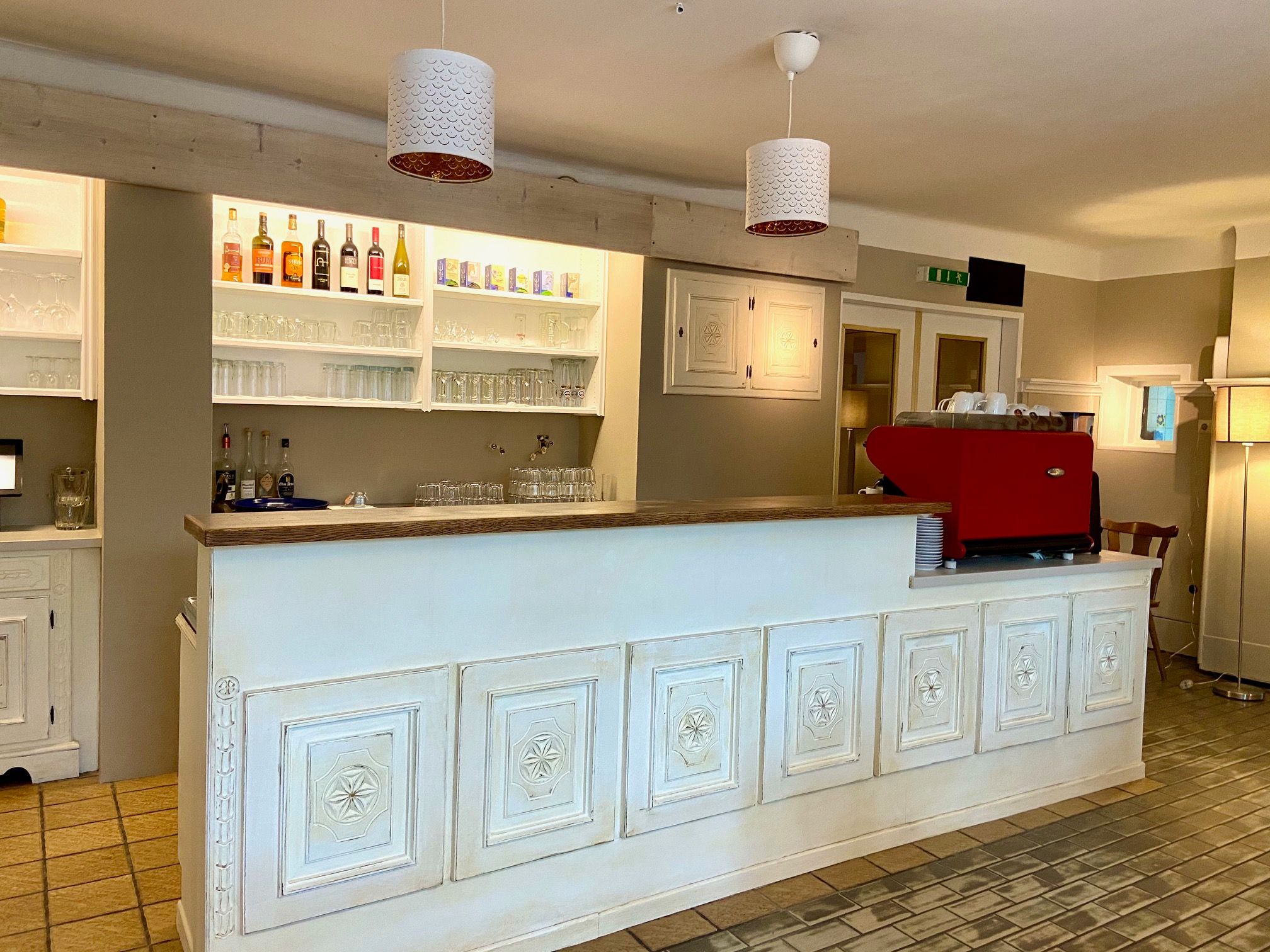 A stylish bar area with a white counter, shelves with bottles and glasses and a red coffee machine.