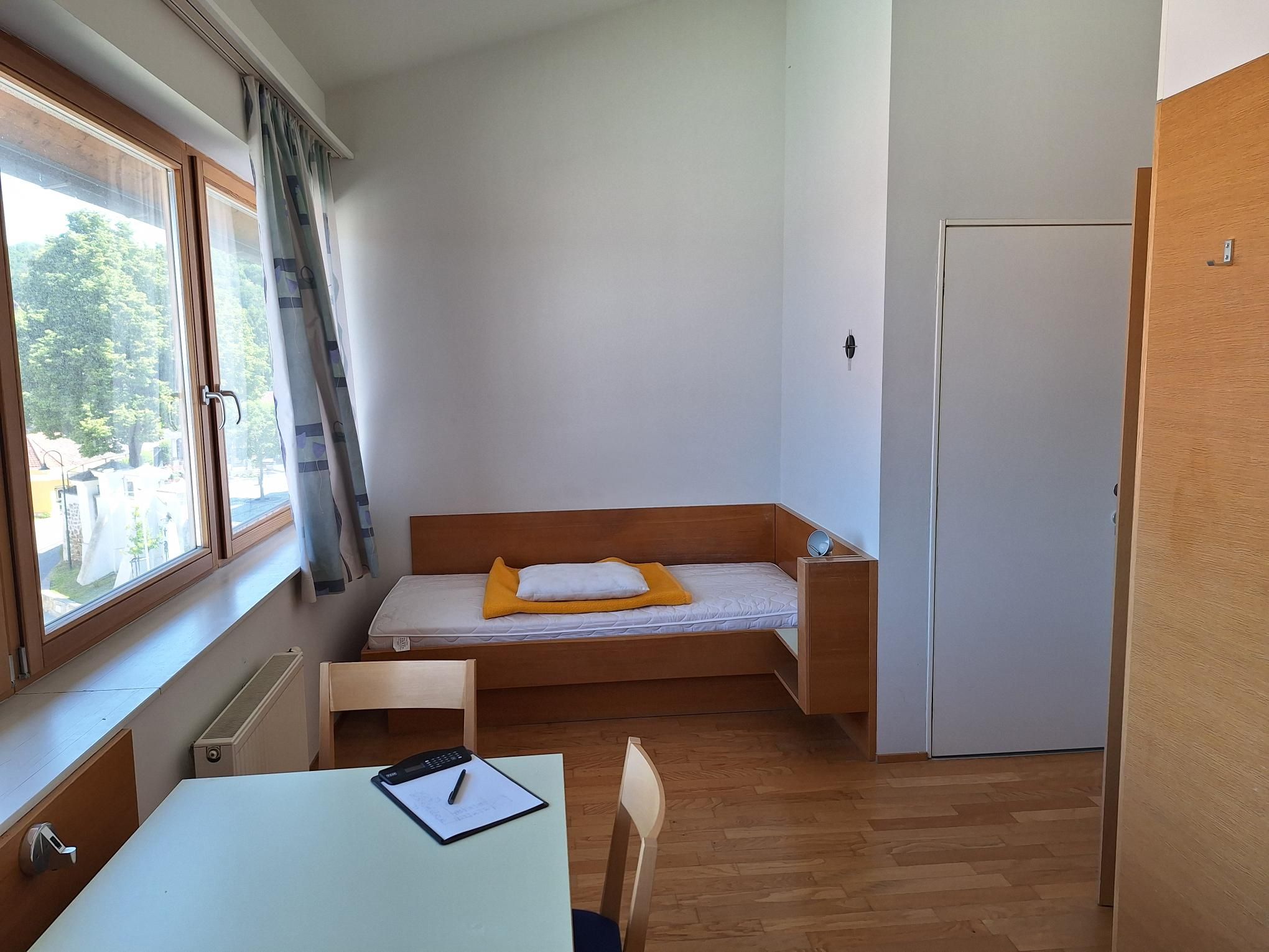 A simply furnished single room with a bed, table and chair, light wooden furniture and a large window with a view outside.