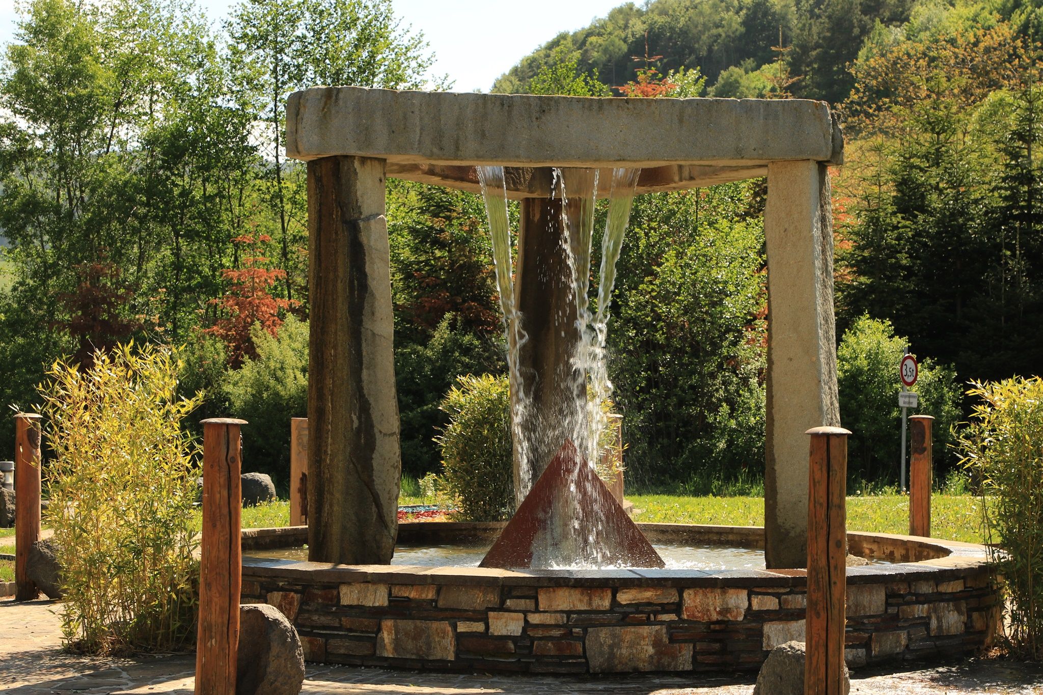 A modern outdoor fountain with a pyramid-shaped structure in the center, surrounded by trees and plants.