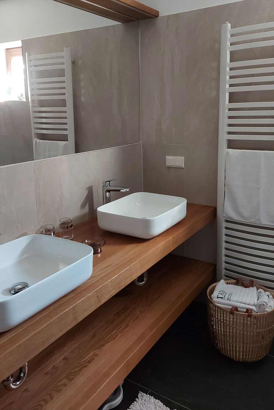 Modern bathroom with two white washbasins on a wooden top, towel rail and basket with towels.