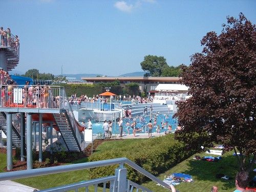 A lively outdoor pool with lots of people, water slides and sunbathing lawn.