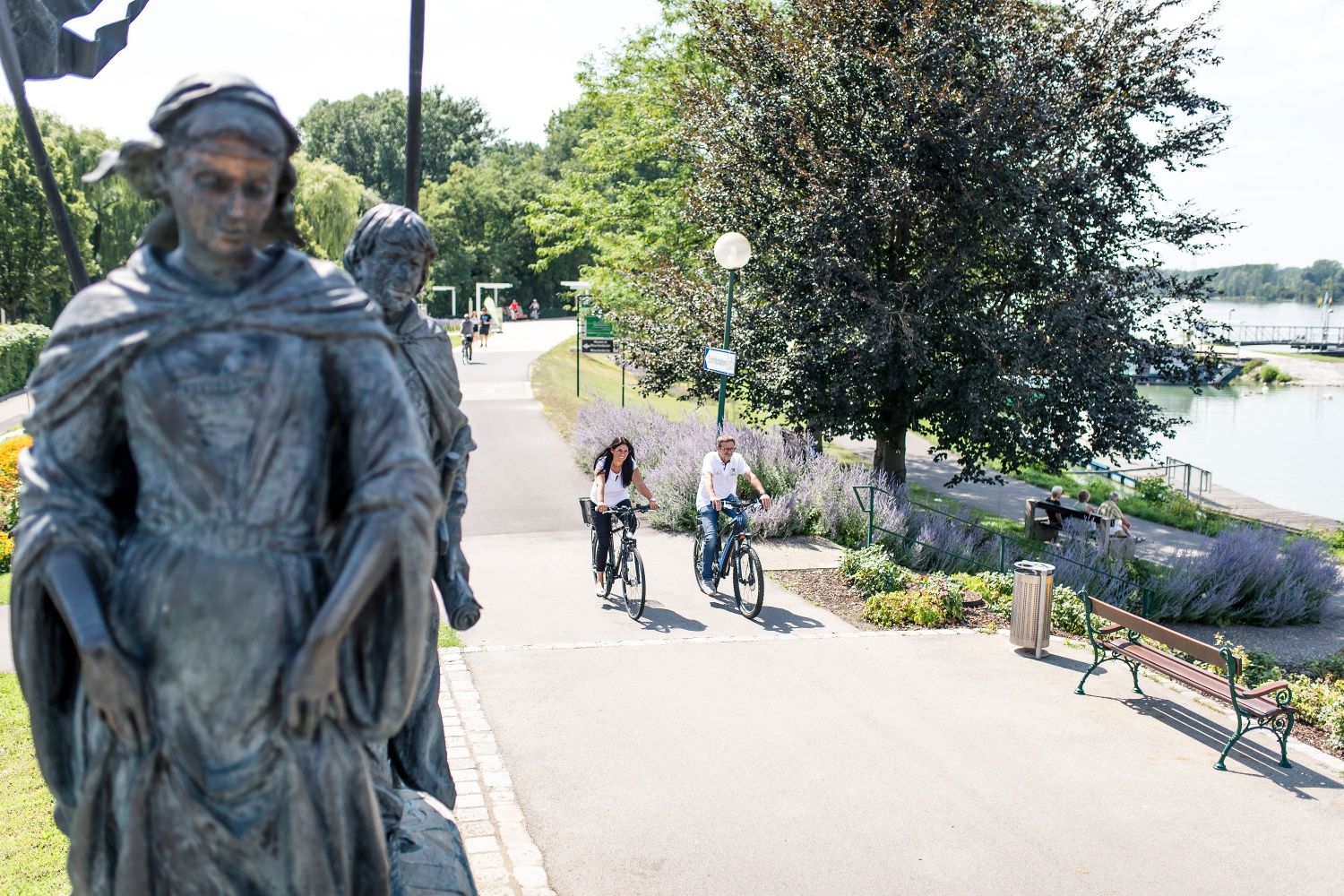 Two people ride past a statue on a cycle path, surrounded by trees and a river in the background.