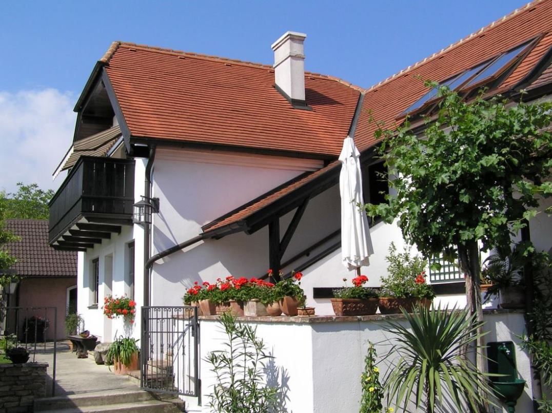 A white house with a red tiled roof, balcony and garden with plants and flowers.