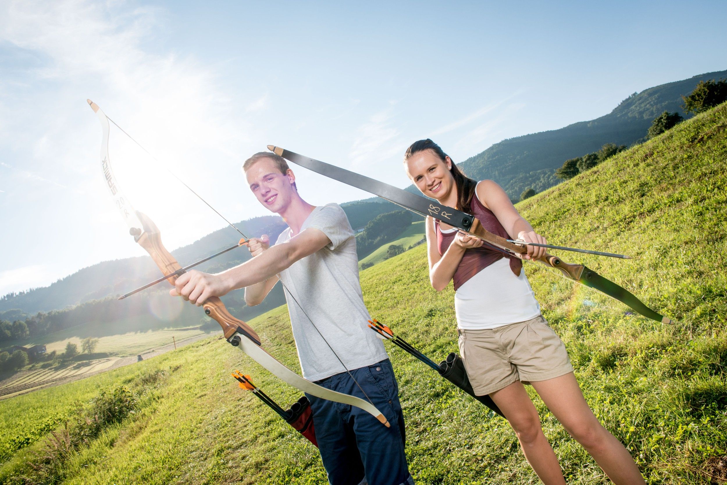 Two people doing archery in a meadow with mountains in the background.