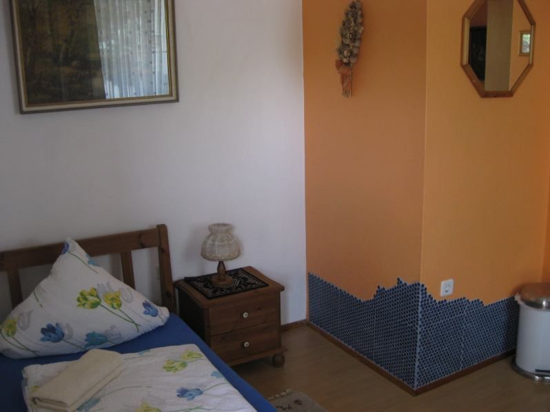 A small room with a single bed, bedside table, lamp and orange wall with mosaic tiles.