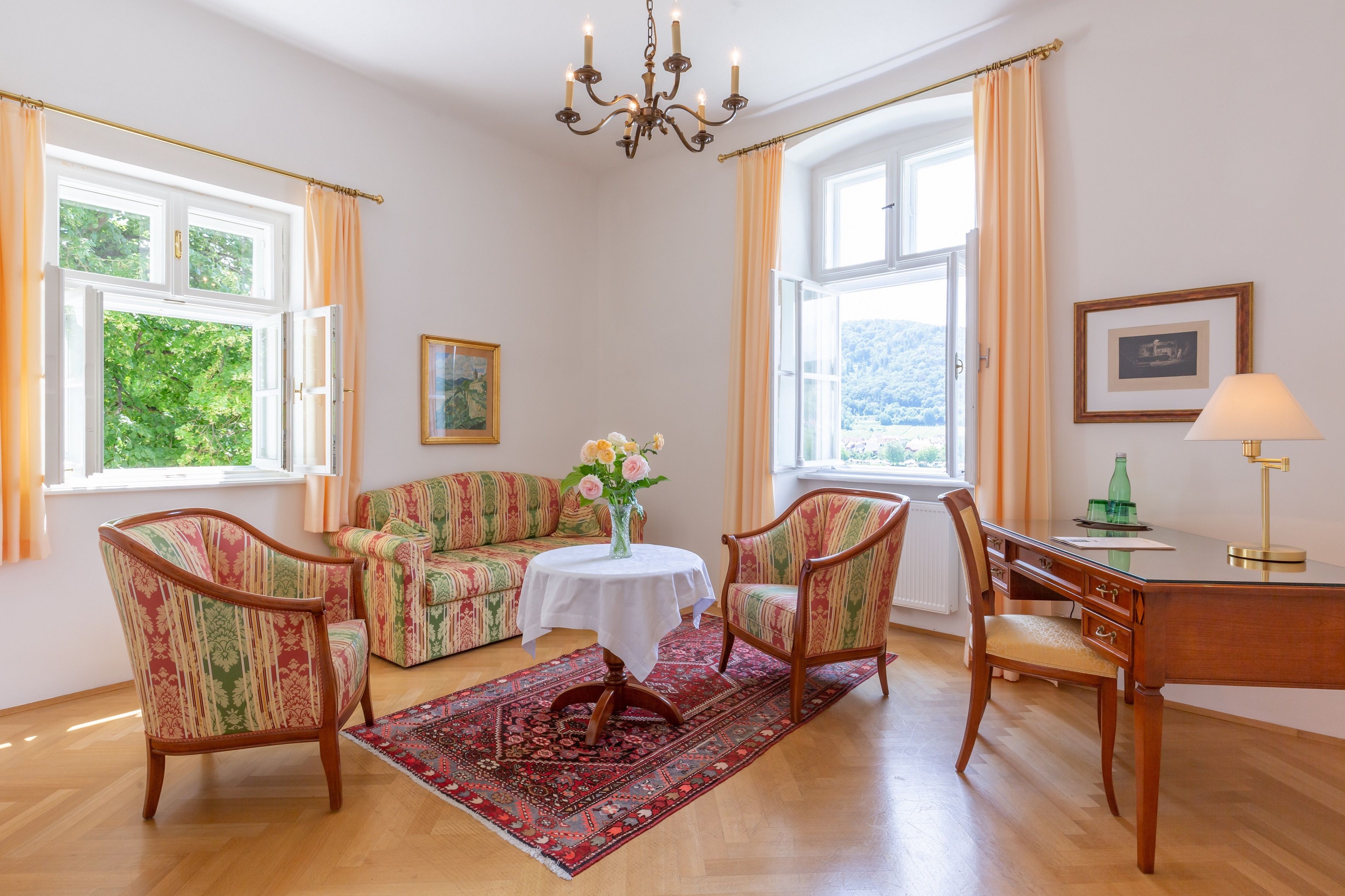 Elegant living room with antique furniture, two windows, a table with flowers, a desk and a carpet on a wooden floor.