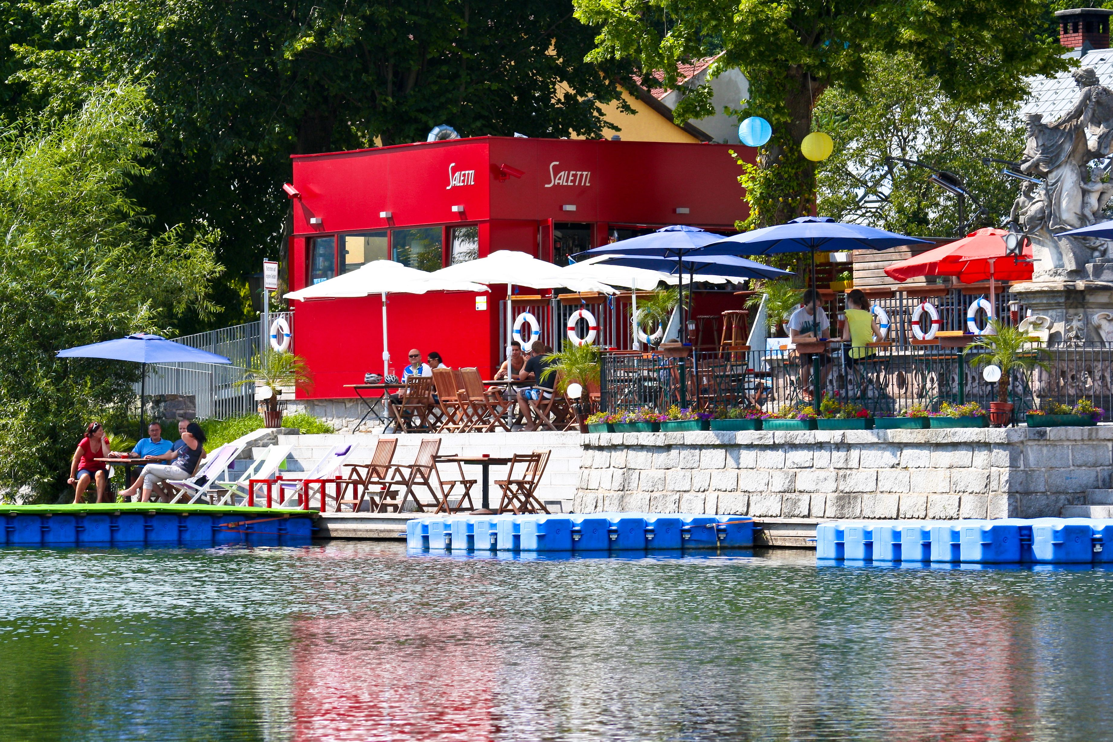 A red building with a terrace by the water, surrounded by parasols and seating.