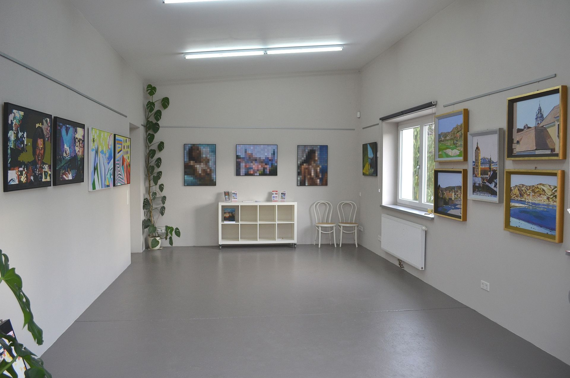 Interior view of a gallery with paintings on the walls and a shelf in the middle.