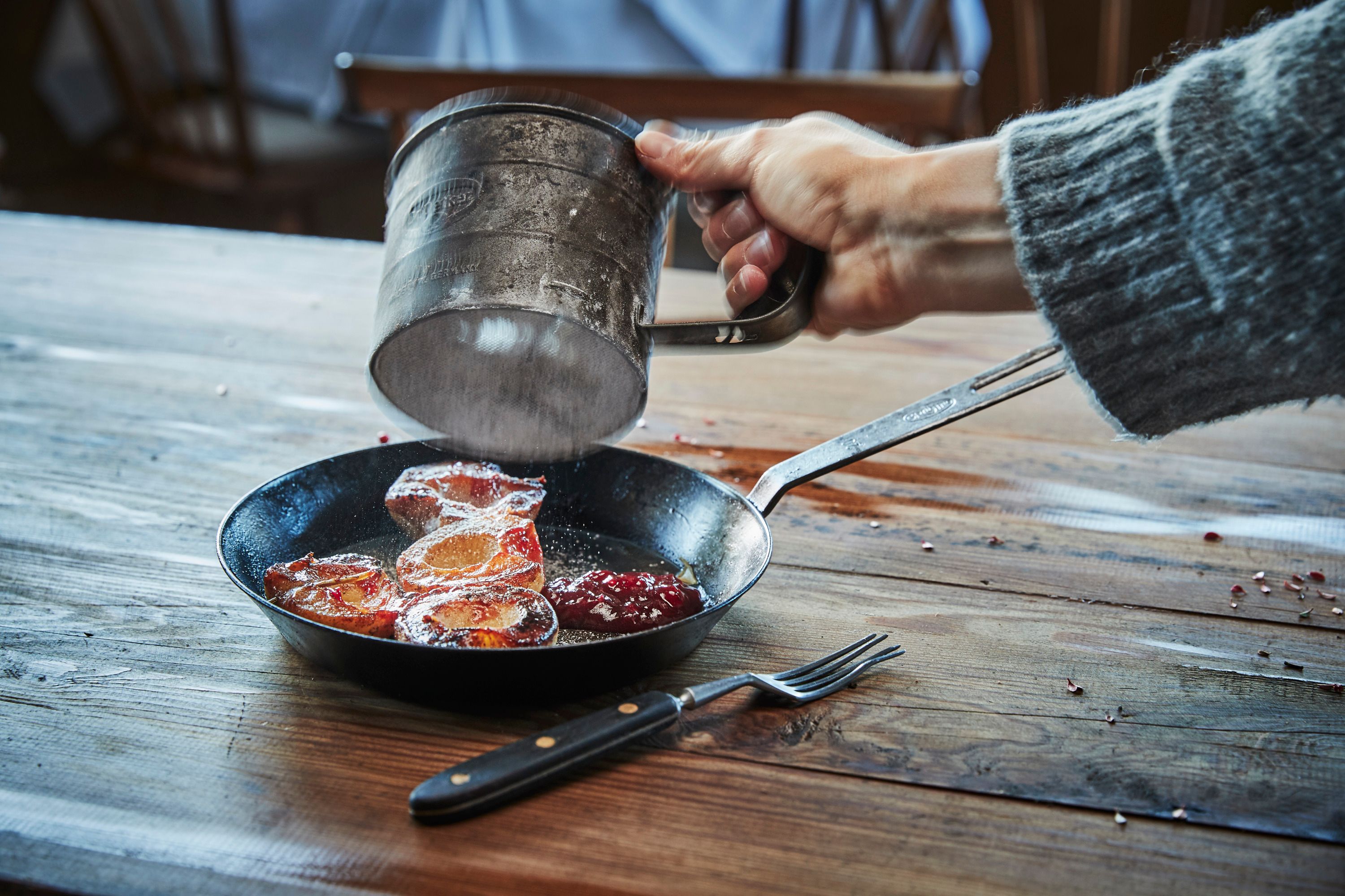 A hand sprinkles powdered sugar over caramelized apple rings in a pan on a wooden table.