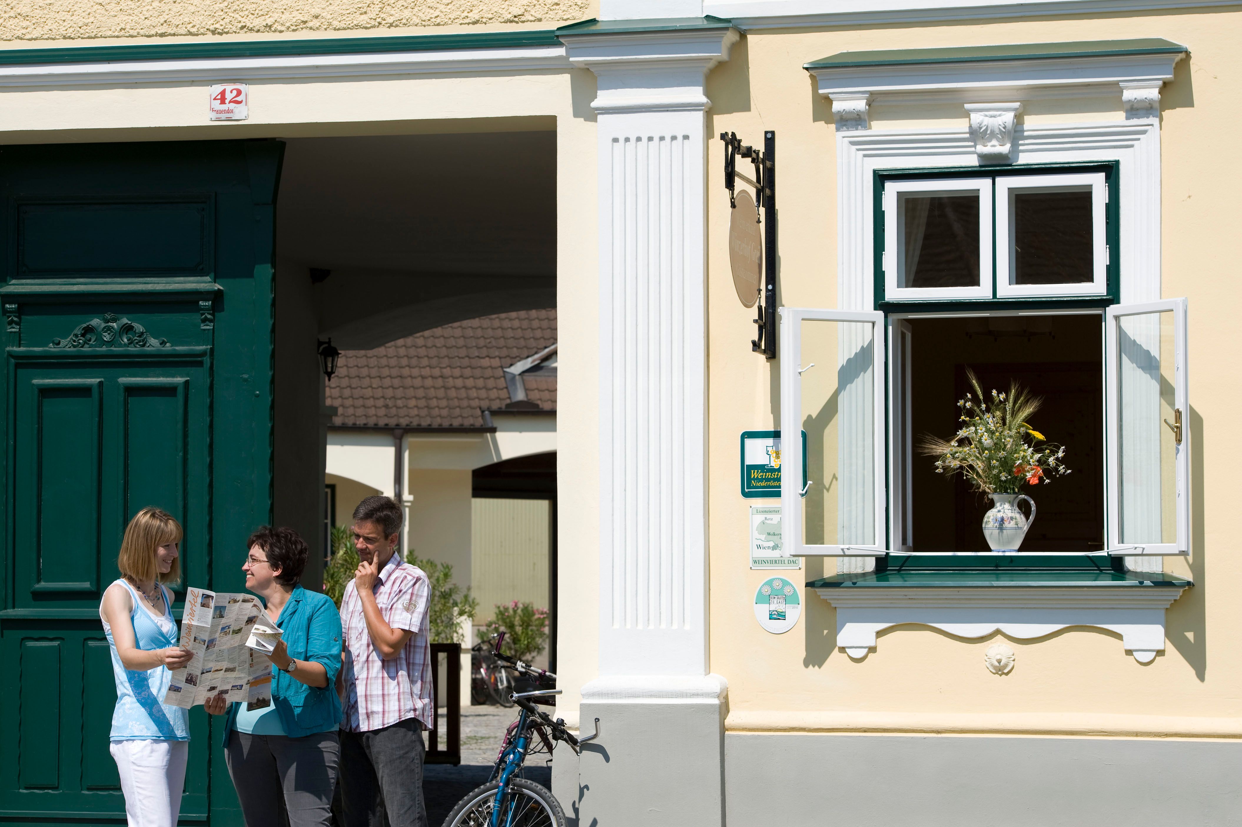 Three people are standing in front of a yellow building with a green door, reading a card. A window with a bouquet of flowers is open.