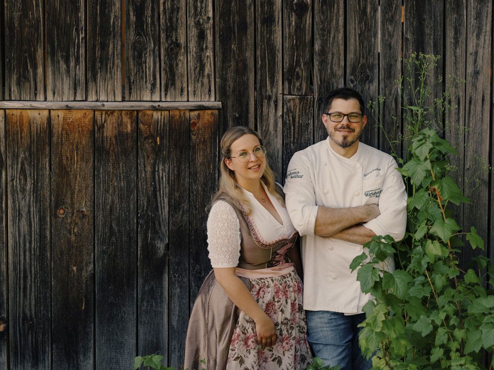 A couple is standing in front of a wooden wall, she is wearing a dirndl, he a chef's jacket.