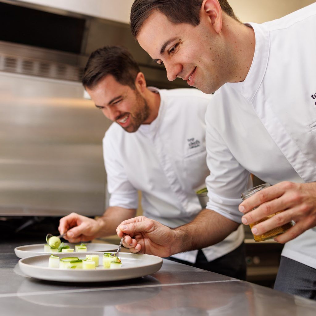 Two chefs in white chef's jackets prepare gourmet dishes on white plates.