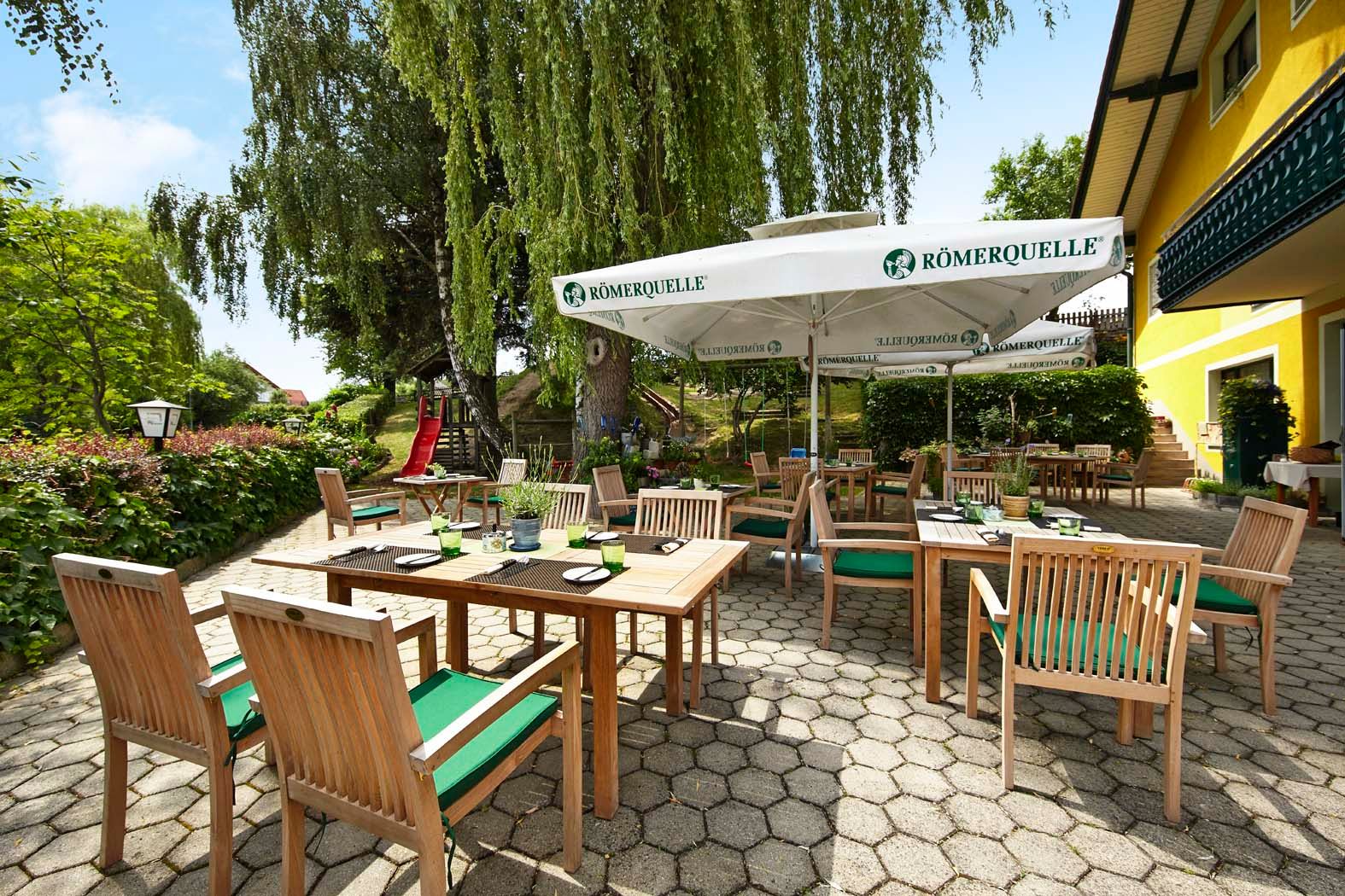 A sunny guest garden with wooden furniture and parasols, surrounded by green vegetation and a yellow building.