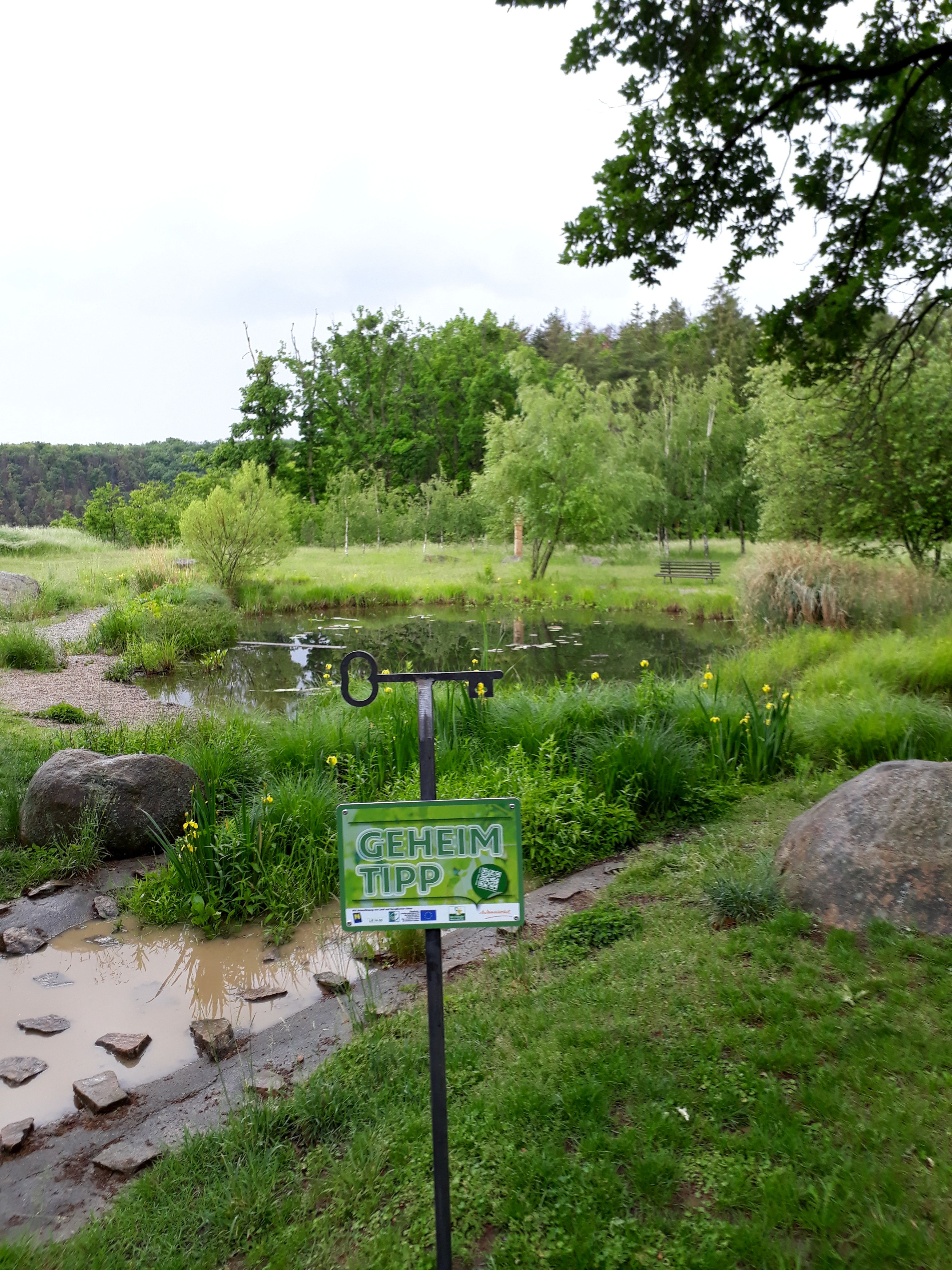 A green pond with trees in the background and a sign saying 'Geheim Tipp' in the foreground.