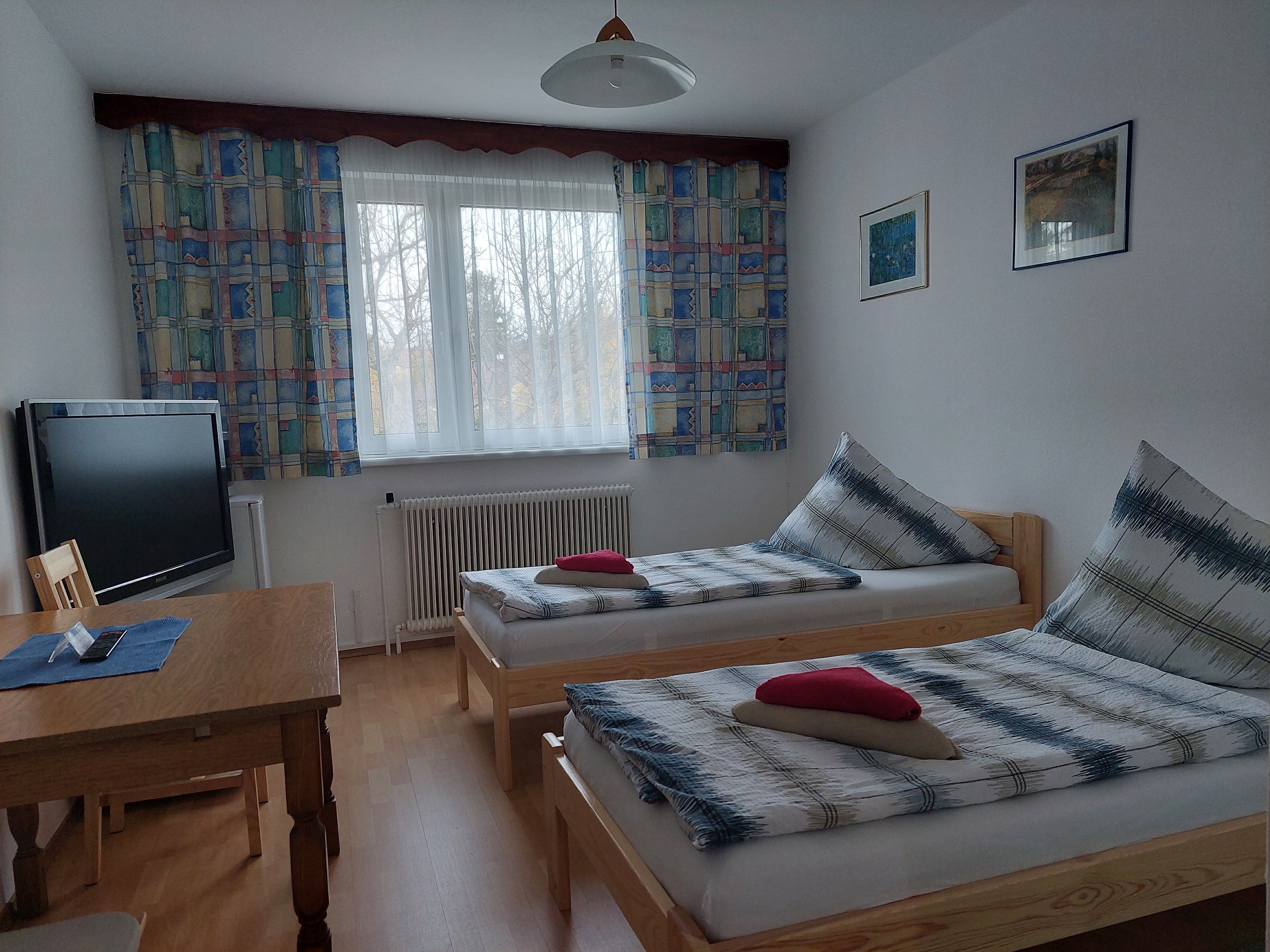 A simply furnished room with two single beds, a table, a TV and a window with colorful curtains.