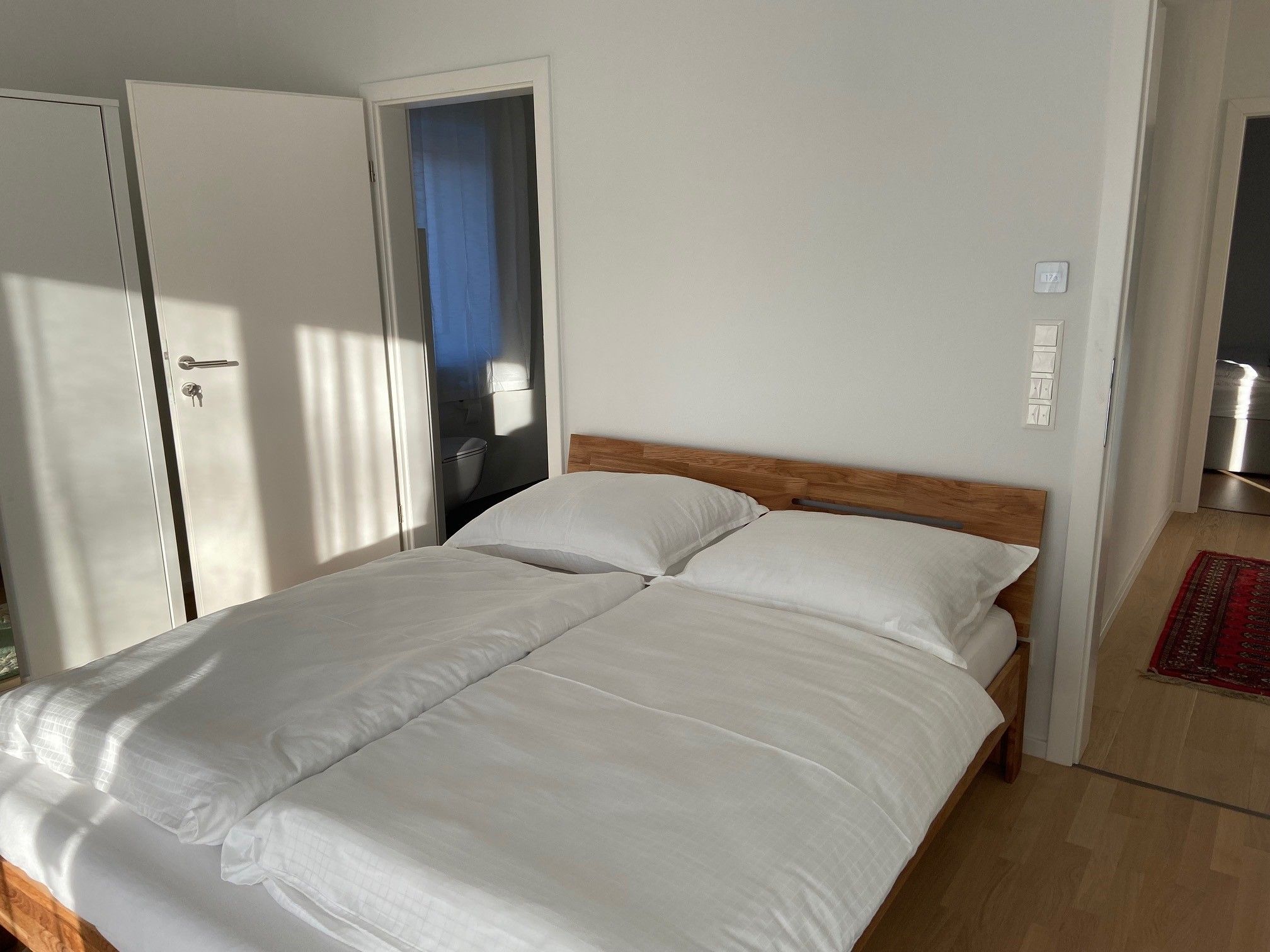 One bedroom with a double bed, white comforter covers and wooden floor. An open door leads to a bathroom.