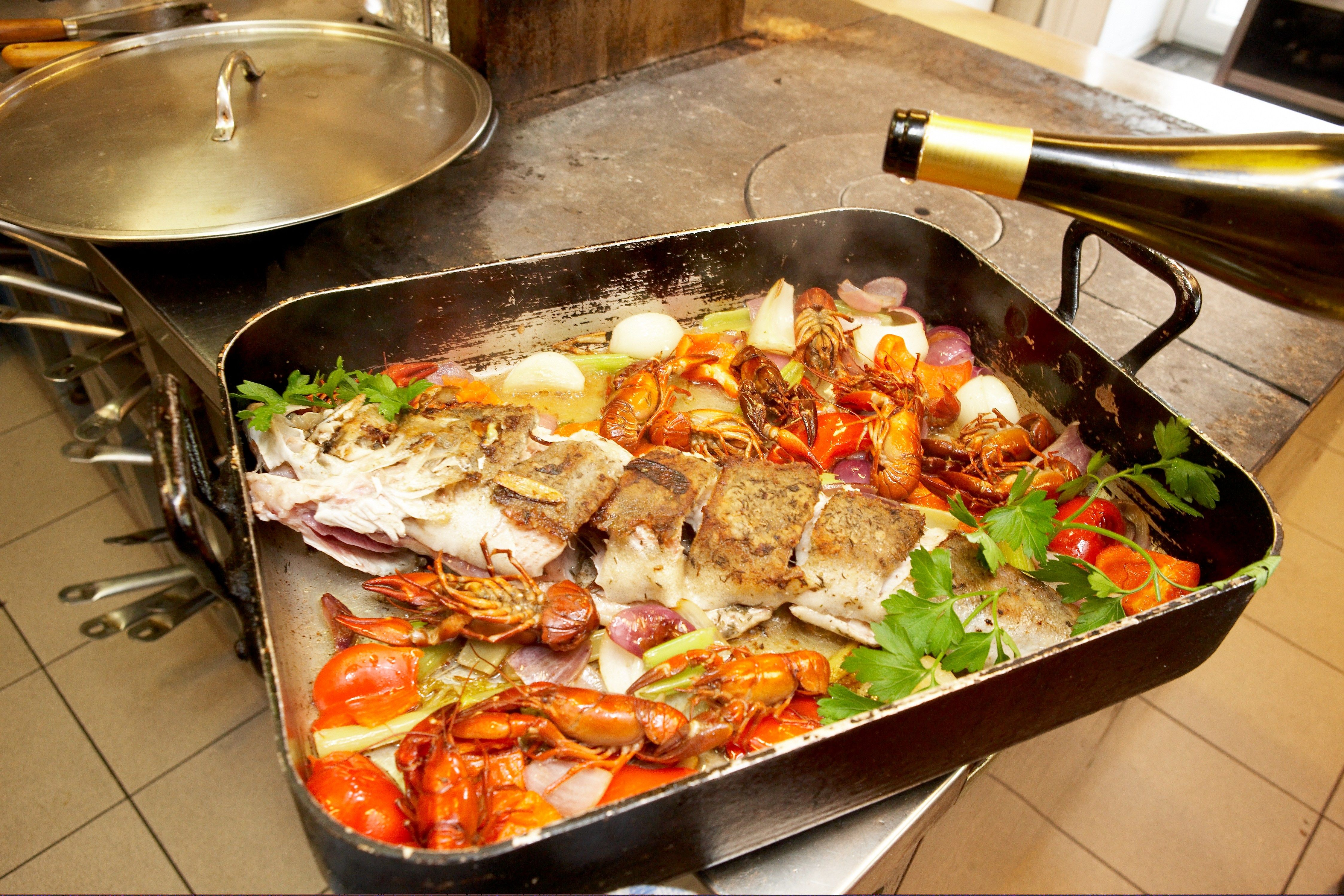 A roasting pan with fish, crayfish, vegetables and herbs on a stove.