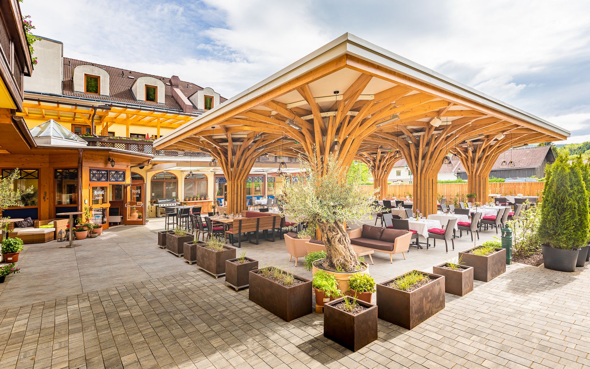 Terrace with wooden roof and tables, surrounded by plants.