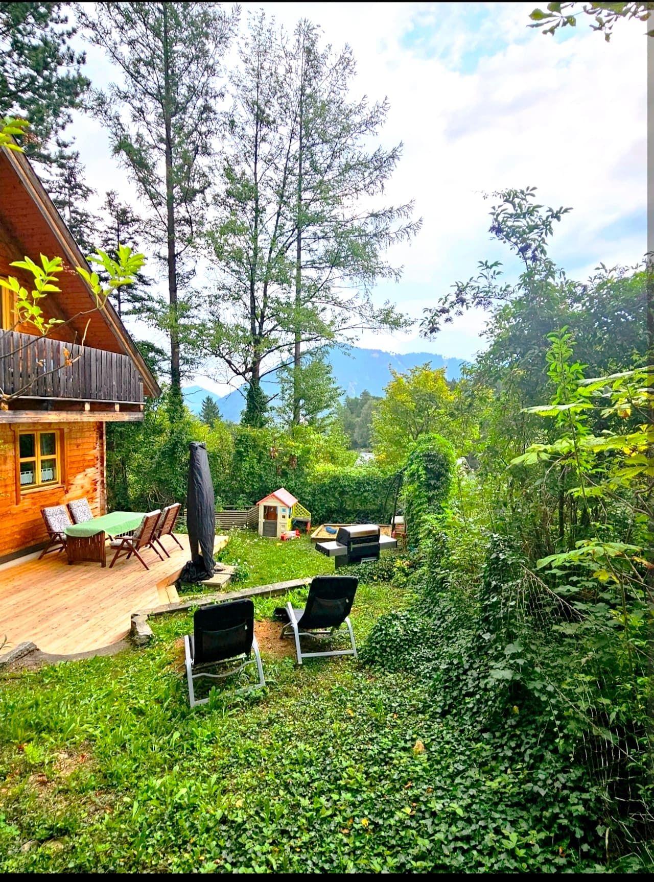 A vacation home with a terrace, garden furniture and sun loungers in a green garden with trees and a view of the mountains.