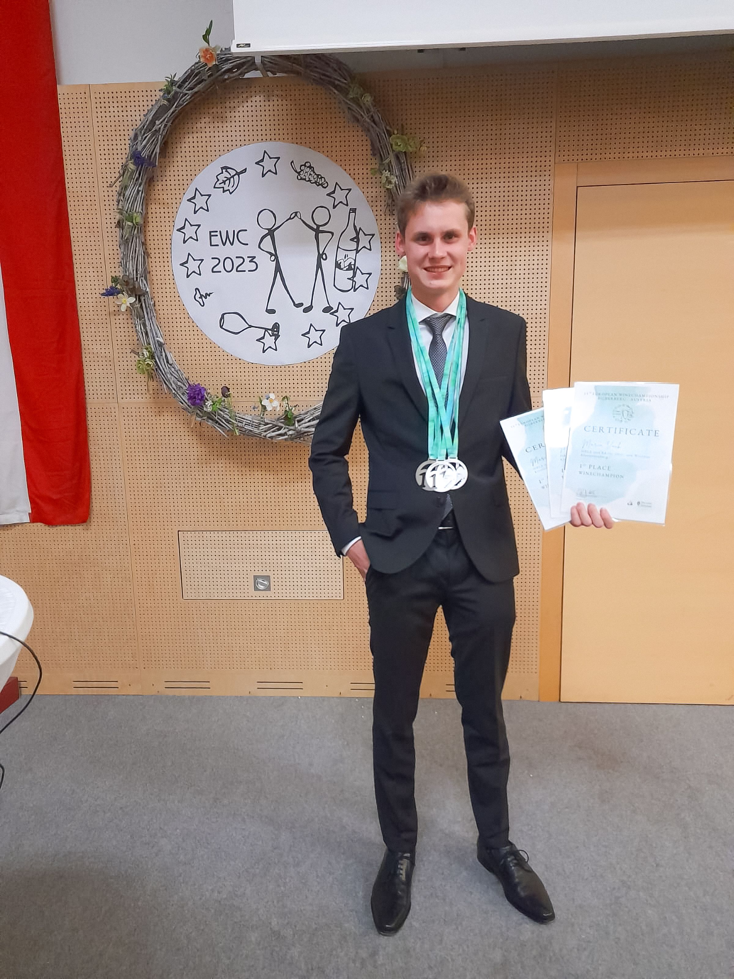 A young man in a suit stands in front of a decorative wreath with a certificate and medals in his hand.