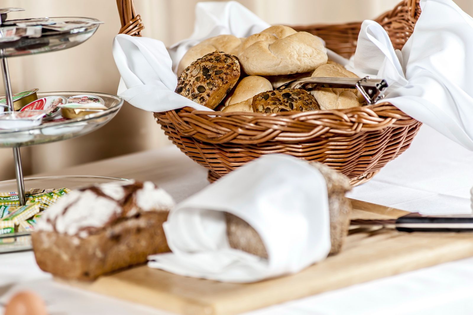 A breakfast table with a basket full of bread rolls, a bread cutting board and a tray of jams.
