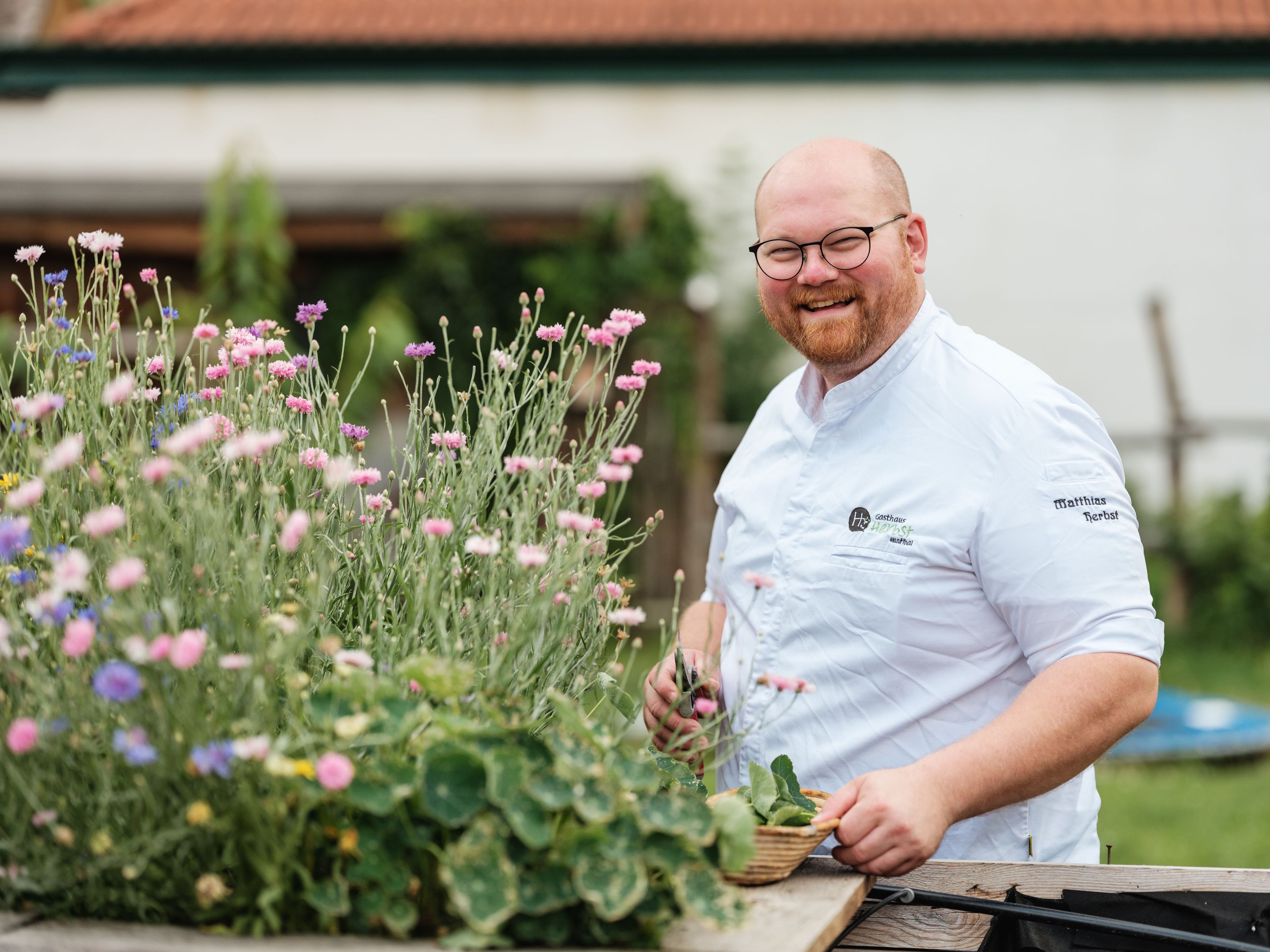 A smiling man in chef's clothes stands next to blooming flowers outside.