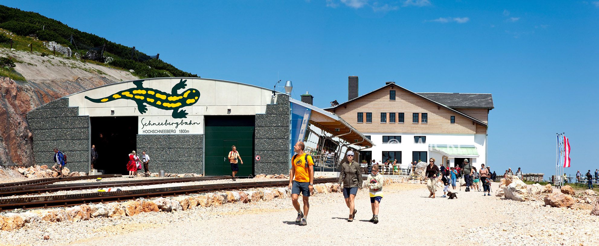 Entrance to the Schneebergbahn on the Hochschneeberg with people and mountain lodge in the background.