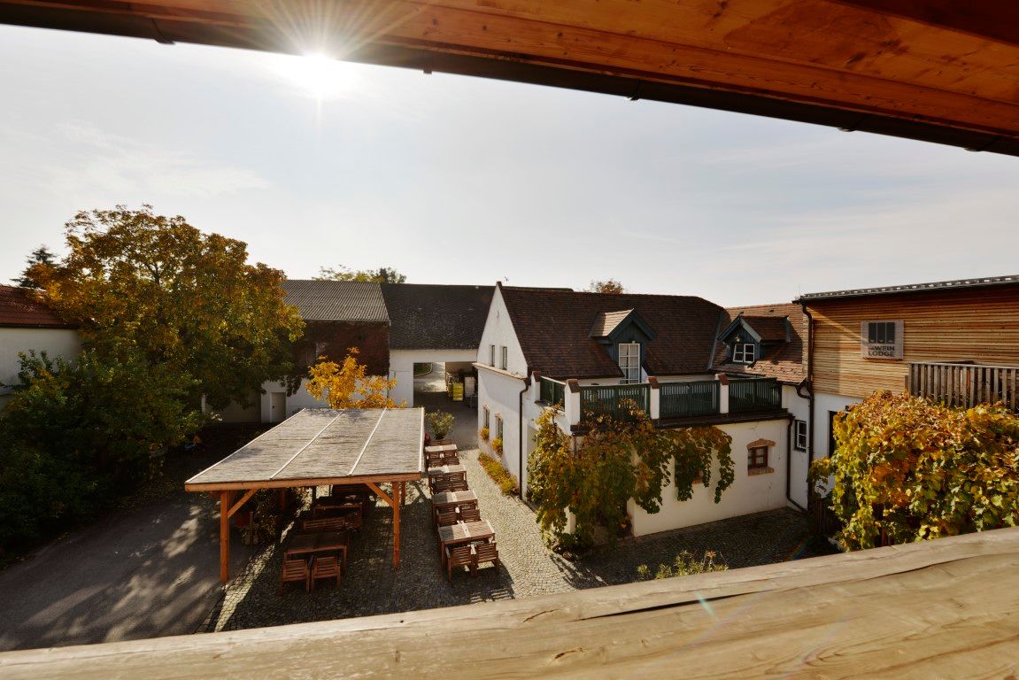 Courtyard of a winery with tables and chairs under a wooden pavilion, surrounded by buildings and autumnal foliage.