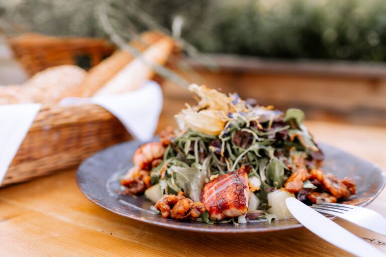 A plate of salad and grilled meat on a wooden table, a basket of bread in the background.