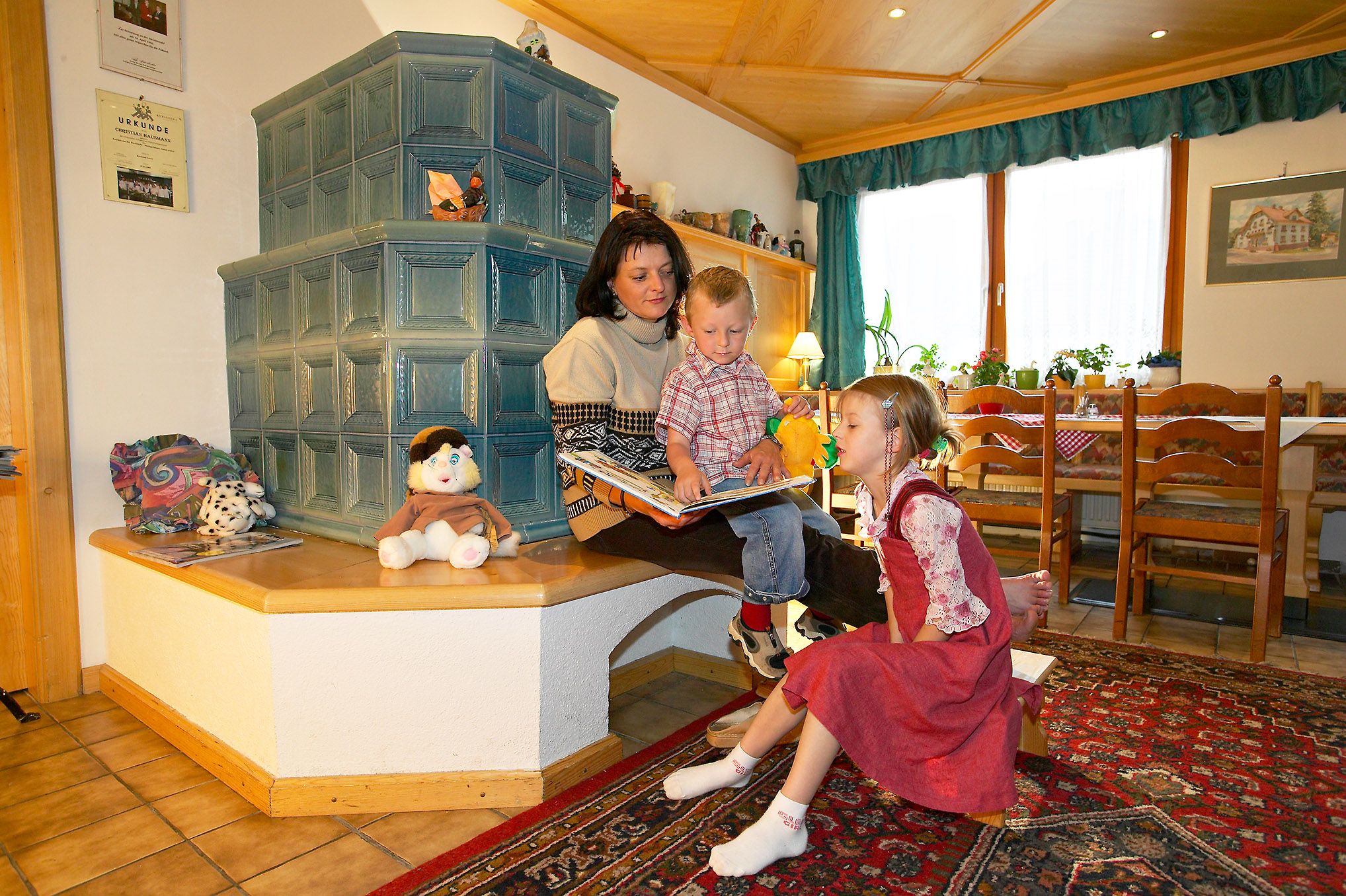 A woman sits with two children on a bench in front of a tiled stove and reads a book. Toys and decorations are scattered around her.